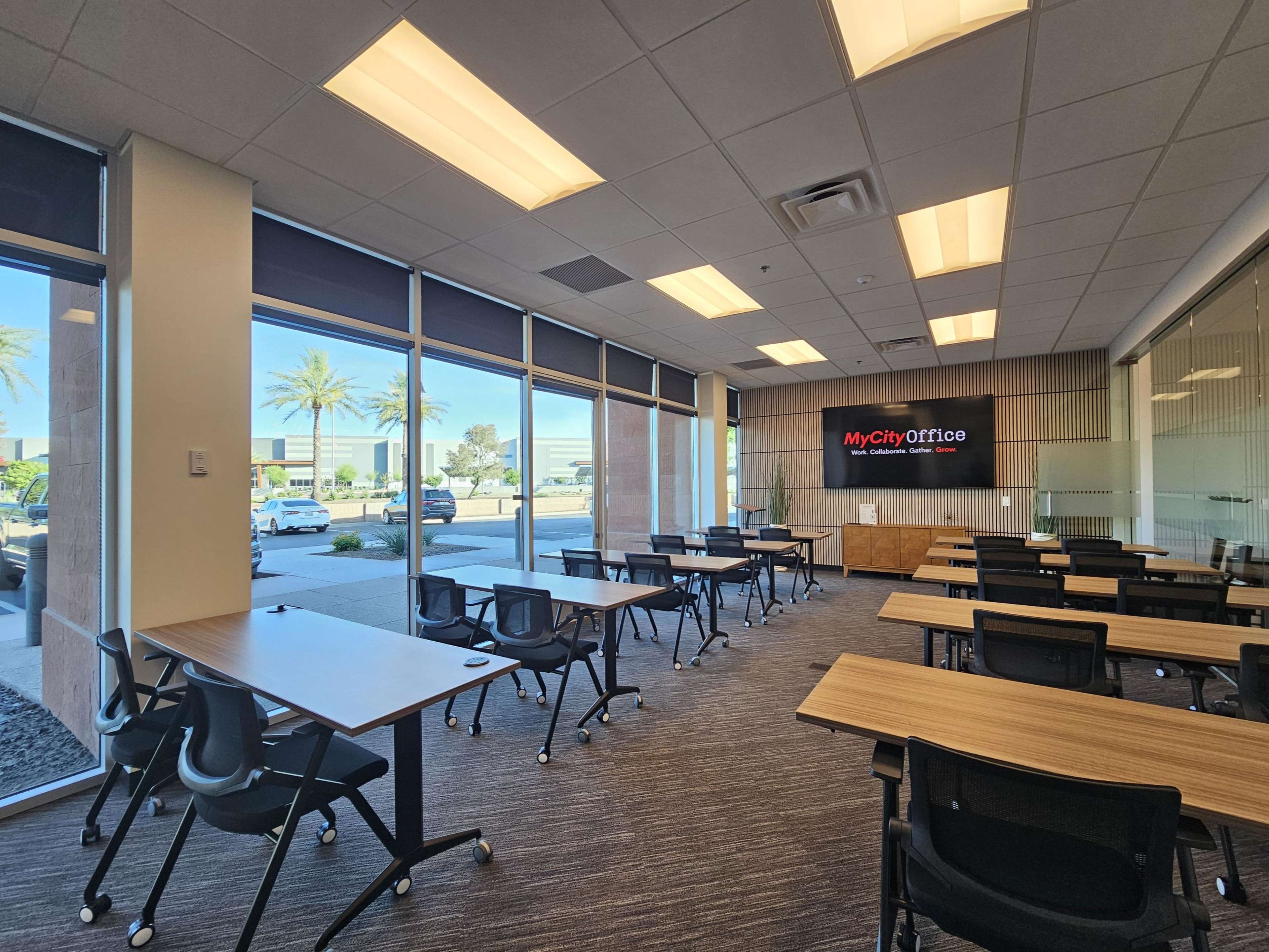The image shows a modern conference room with several tables and chairs arranged for a meeting, large windows letting in natural light, and a display screen showing a logo.