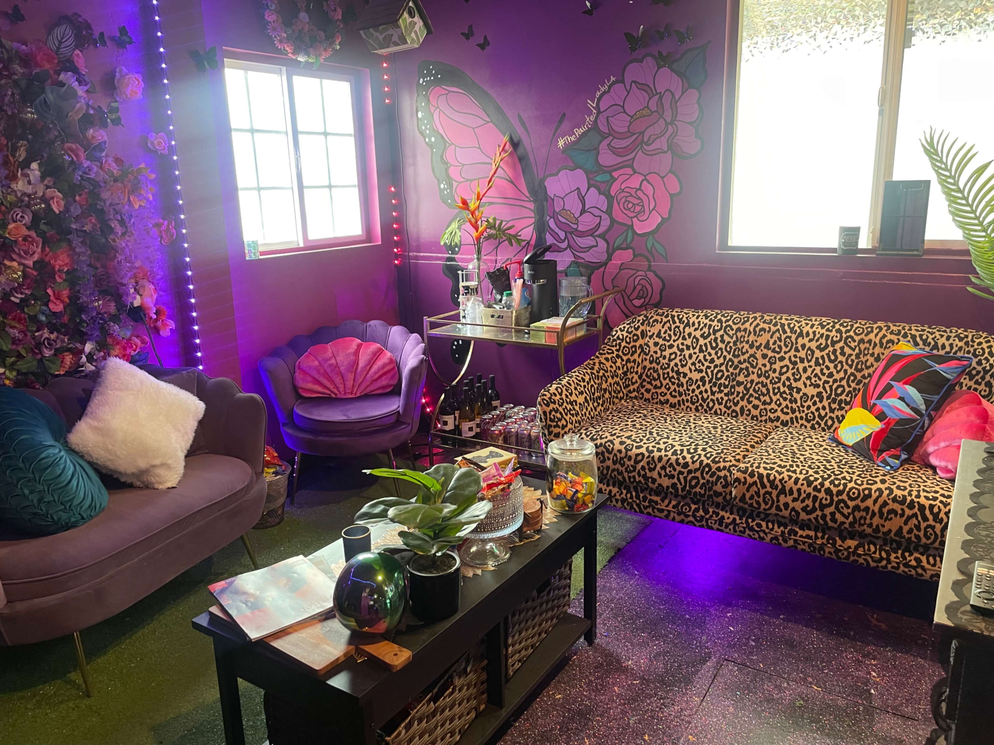 The image shows a colorful room decorated with a floral wall, a leopard print couch, and various lounge furniture around a coffee table.