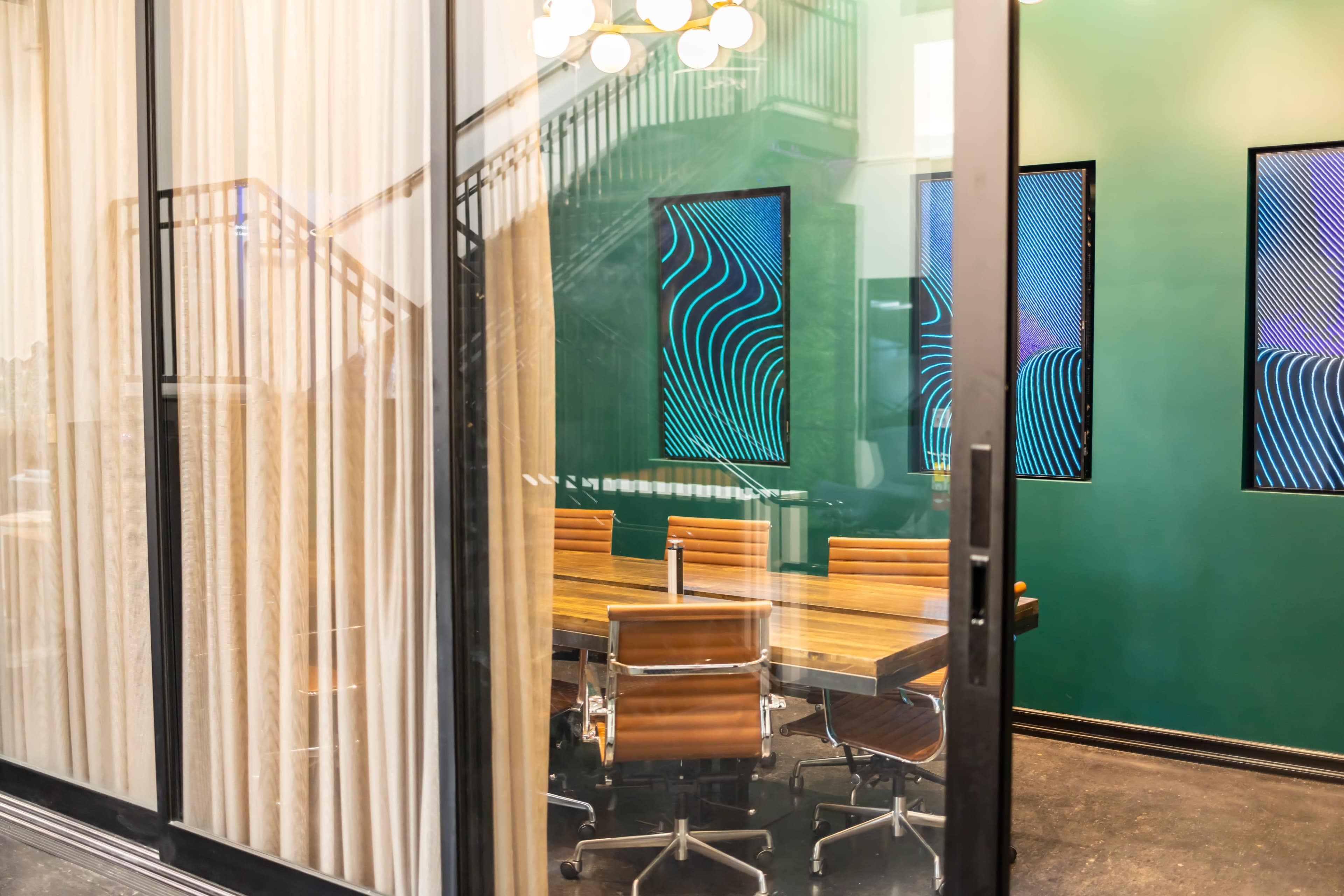 A modern conference room features a wooden table surrounded by leather chairs and displays on the green walls.