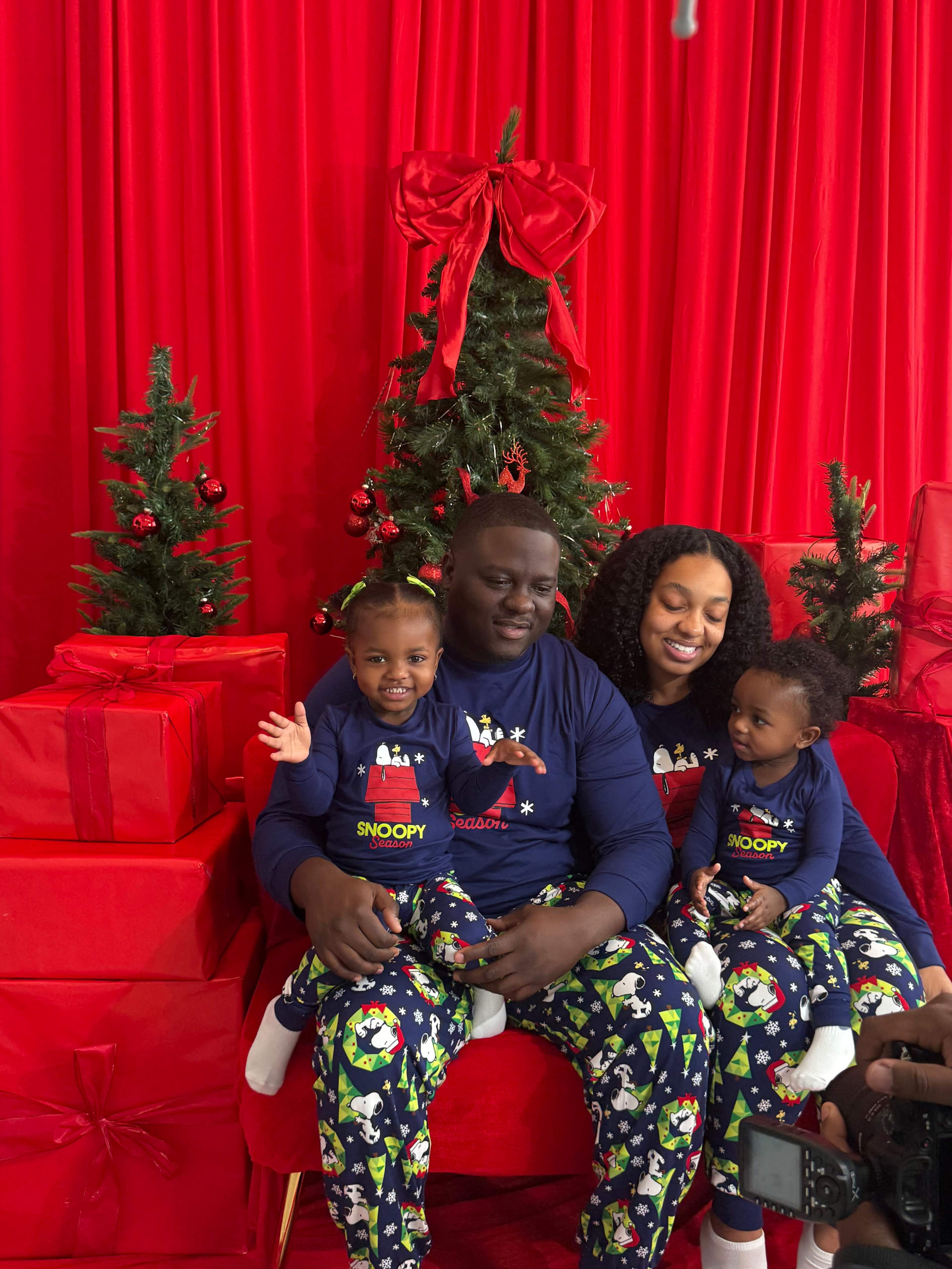 A family sits on a red couch surrounded by Christmas trees and gifts, wearing matching holiday pajamas.