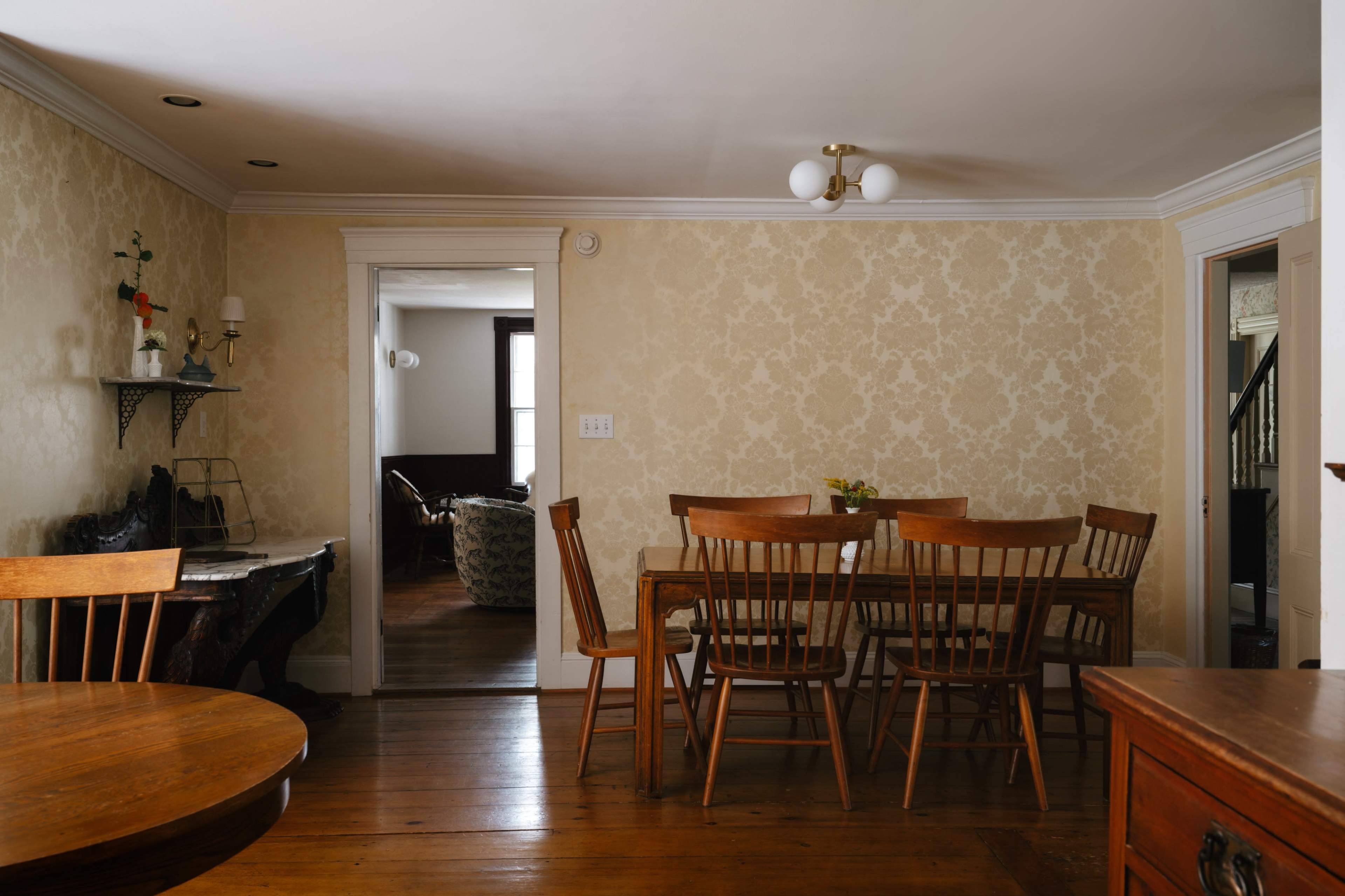 The image shows a dining area with a wooden table and chairs, pale wallpaper, and doorways leading to adjacent rooms.