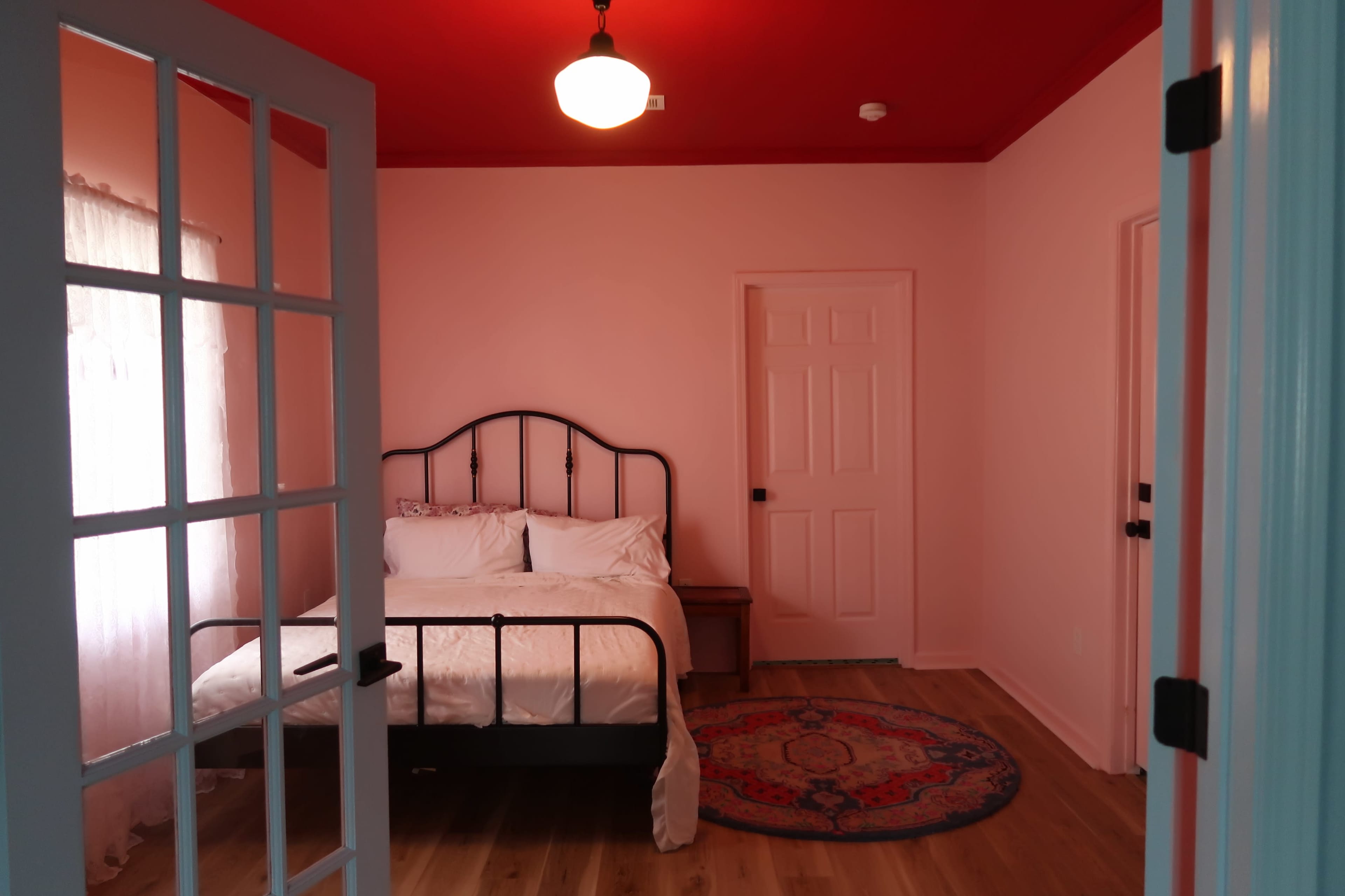 A bedroom with a black metal bed frame, white bedding, a red ceiling, and a wooden floor, viewed through an open door.