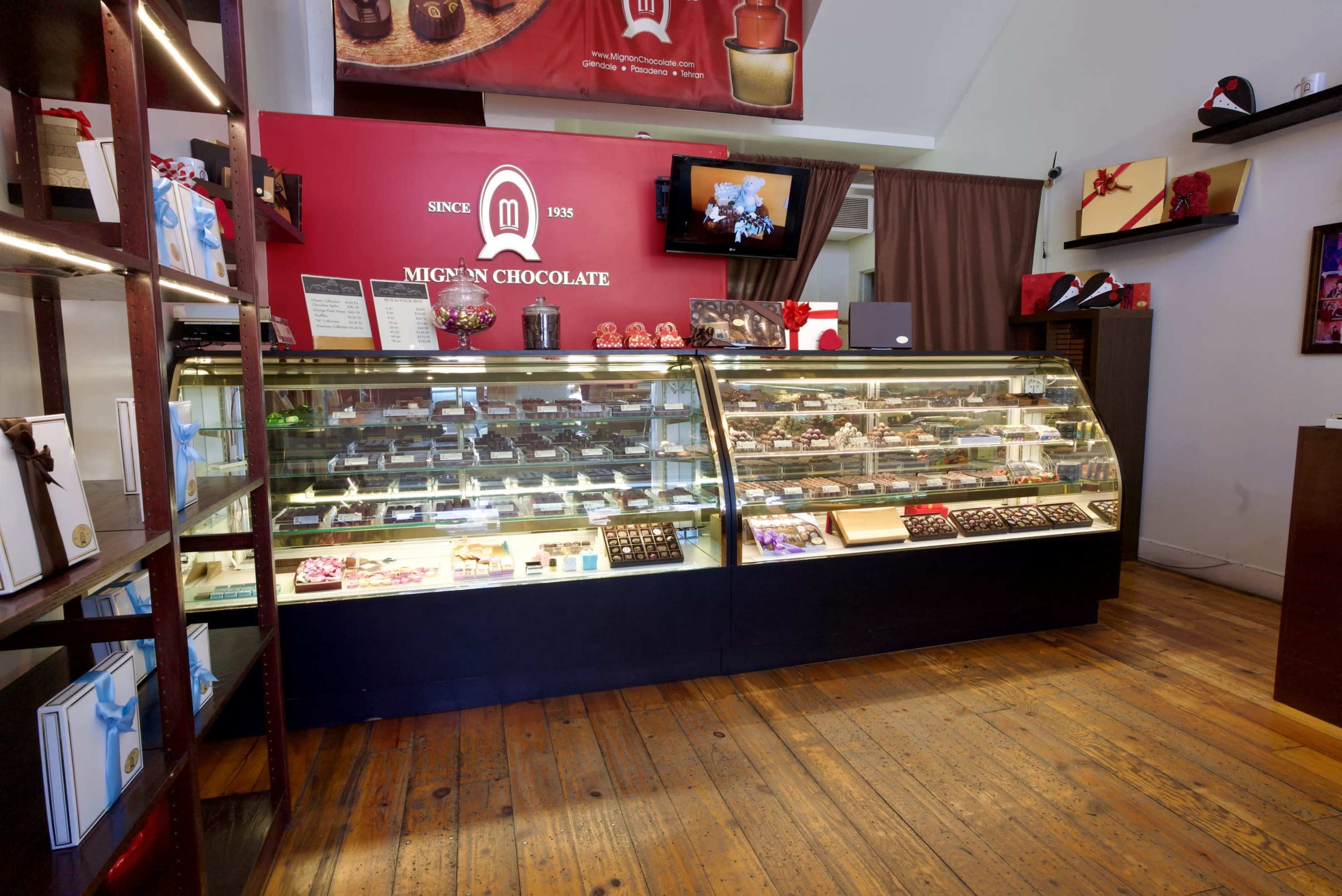 The image shows a chocolate shop with a display case filled with a variety of chocolates, set against a red wall featuring a logo and digital screen.