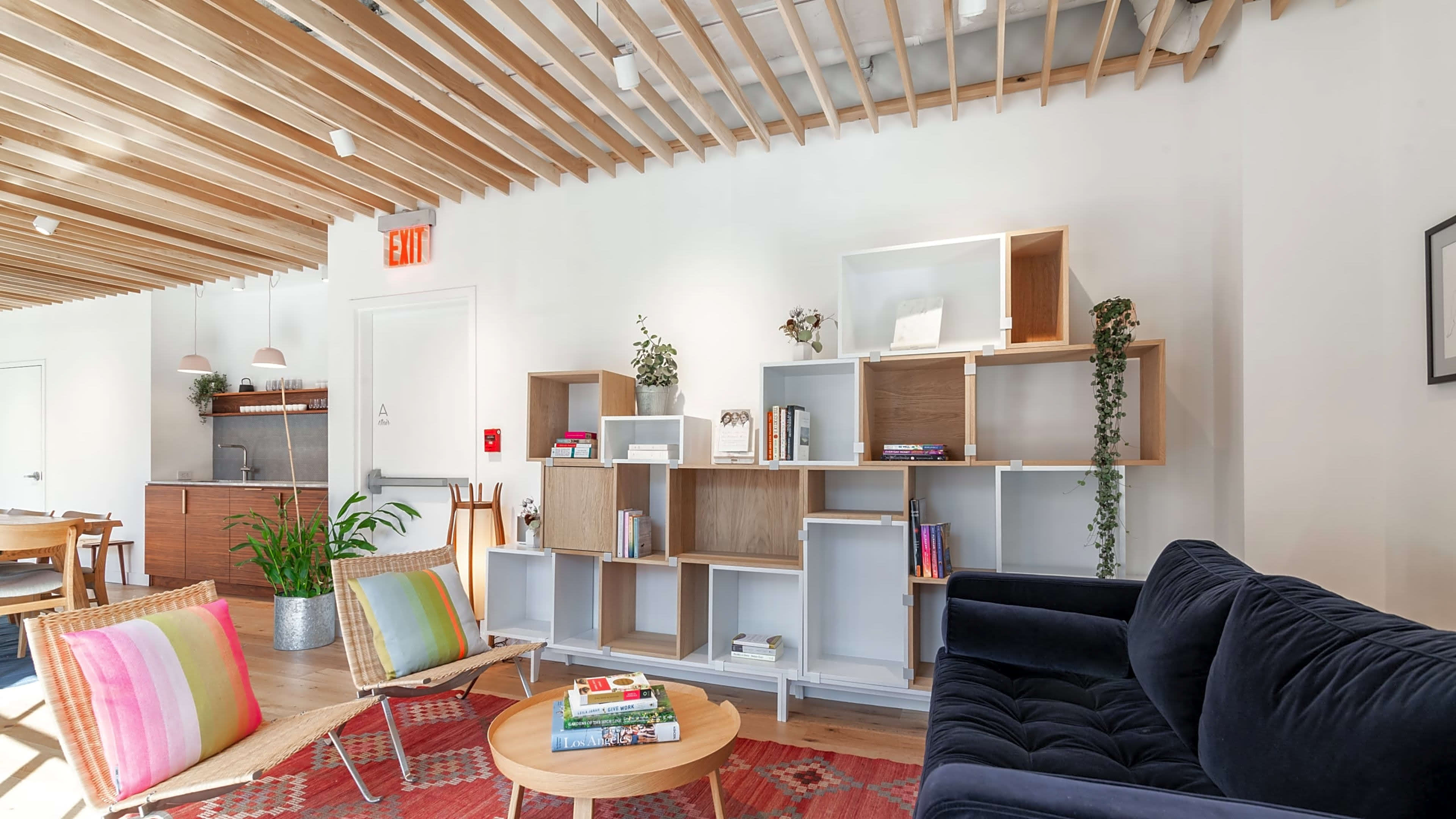 A modern lounge area featuring a navy sofa, a wooden bookshelf with plants and books, and colorful cushions on chairs.