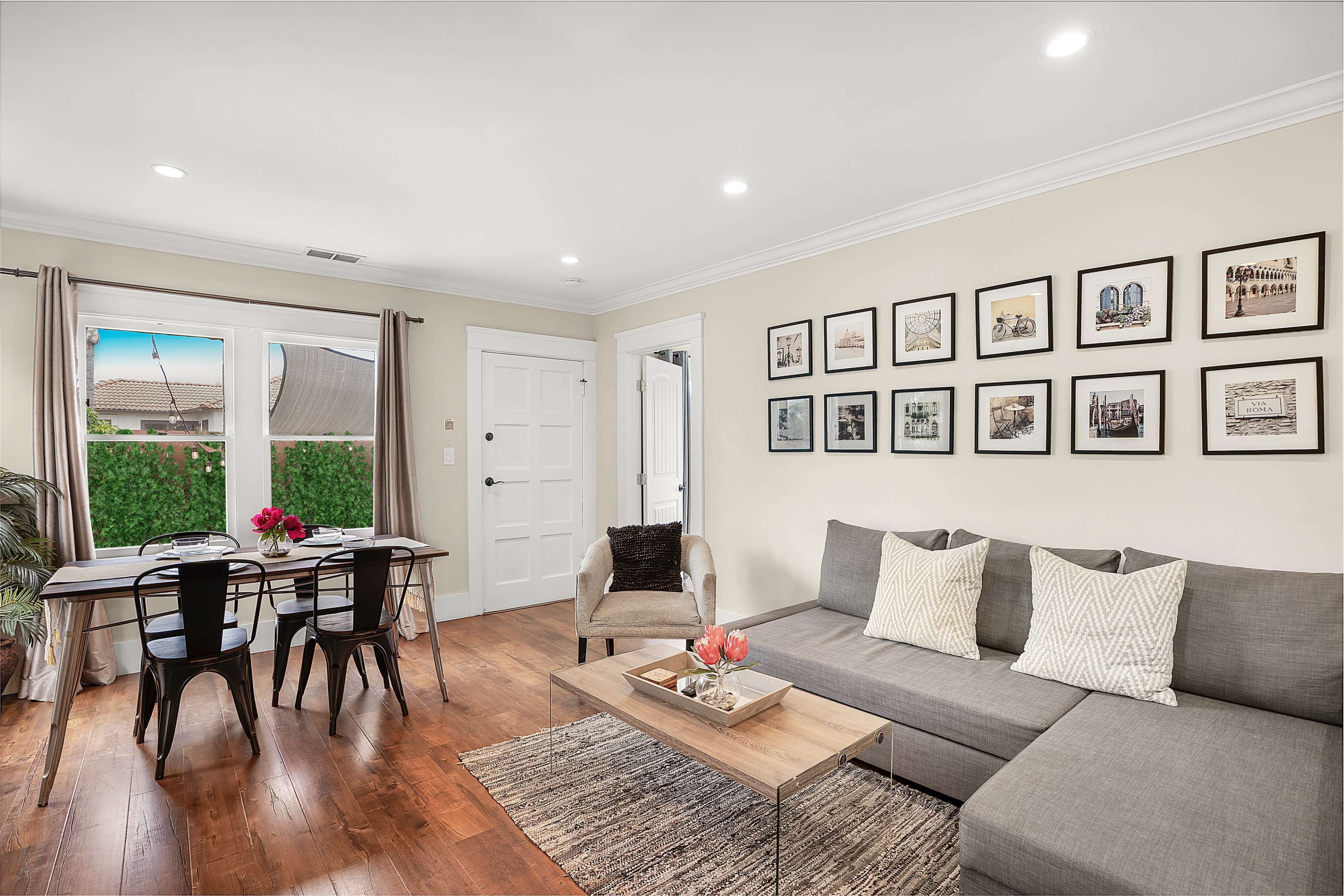 A living room features a sectional sofa, a coffee table with flowers, and a dining area with a table and chairs, all set against a backdrop of framed photographs on the wall.