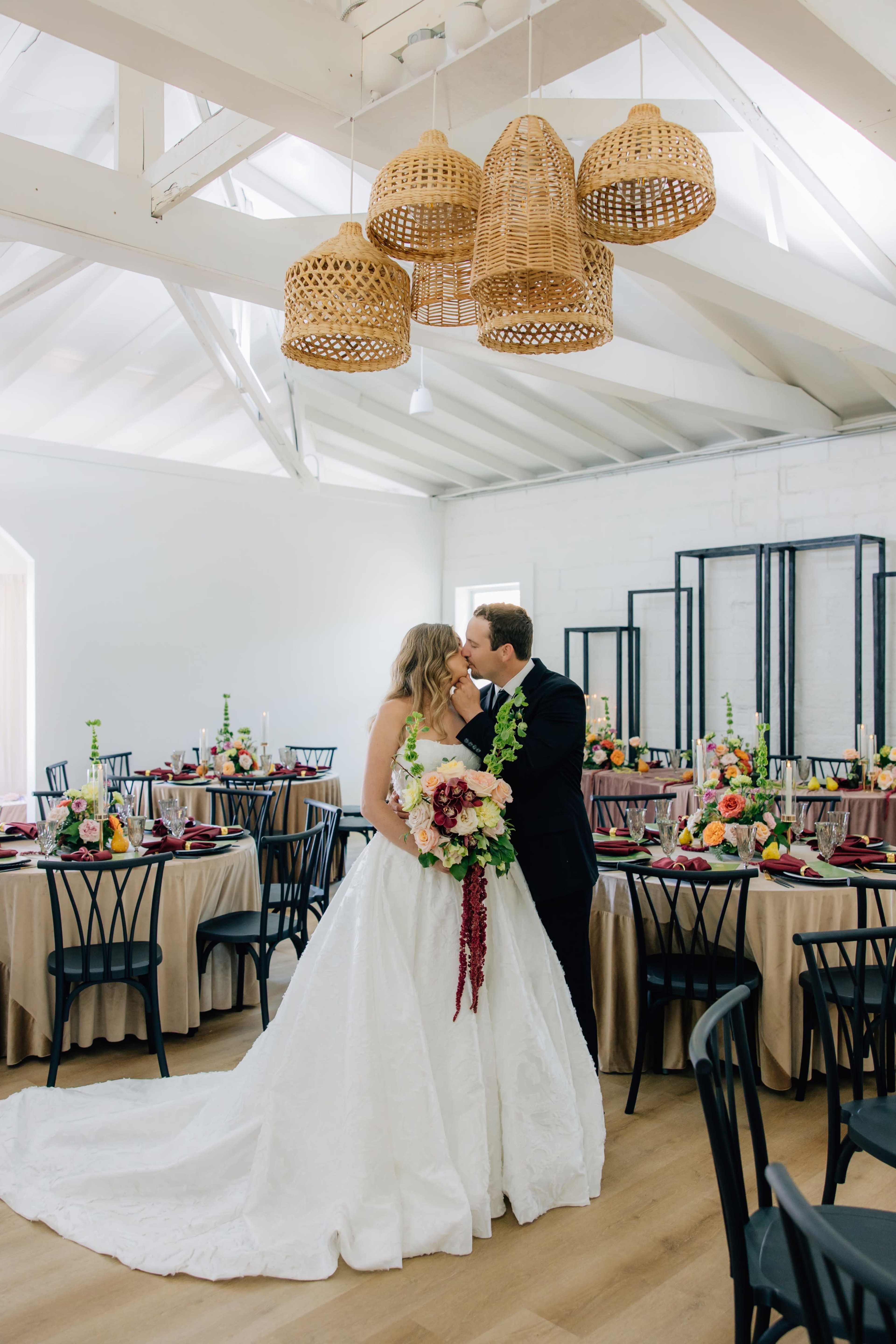 A newlywed couple shares a kiss in a beautifully arranged reception hall adorned with floral centerpieces and decorative lighting.