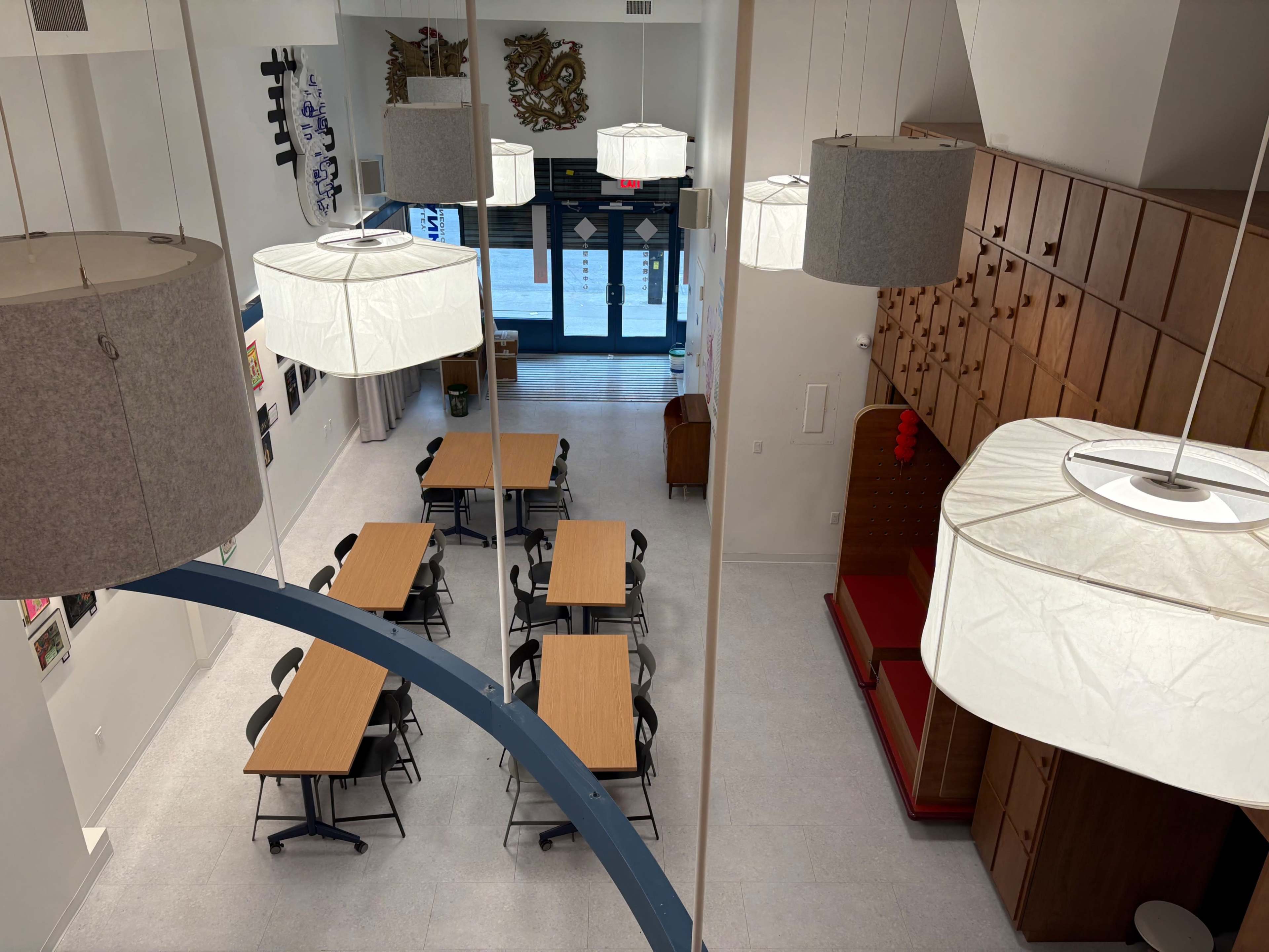 The image shows a spacious, well-lit room with several tables and chairs arranged for seating, large pendant lights hanging from the ceiling, and wooden storage units against one wall.