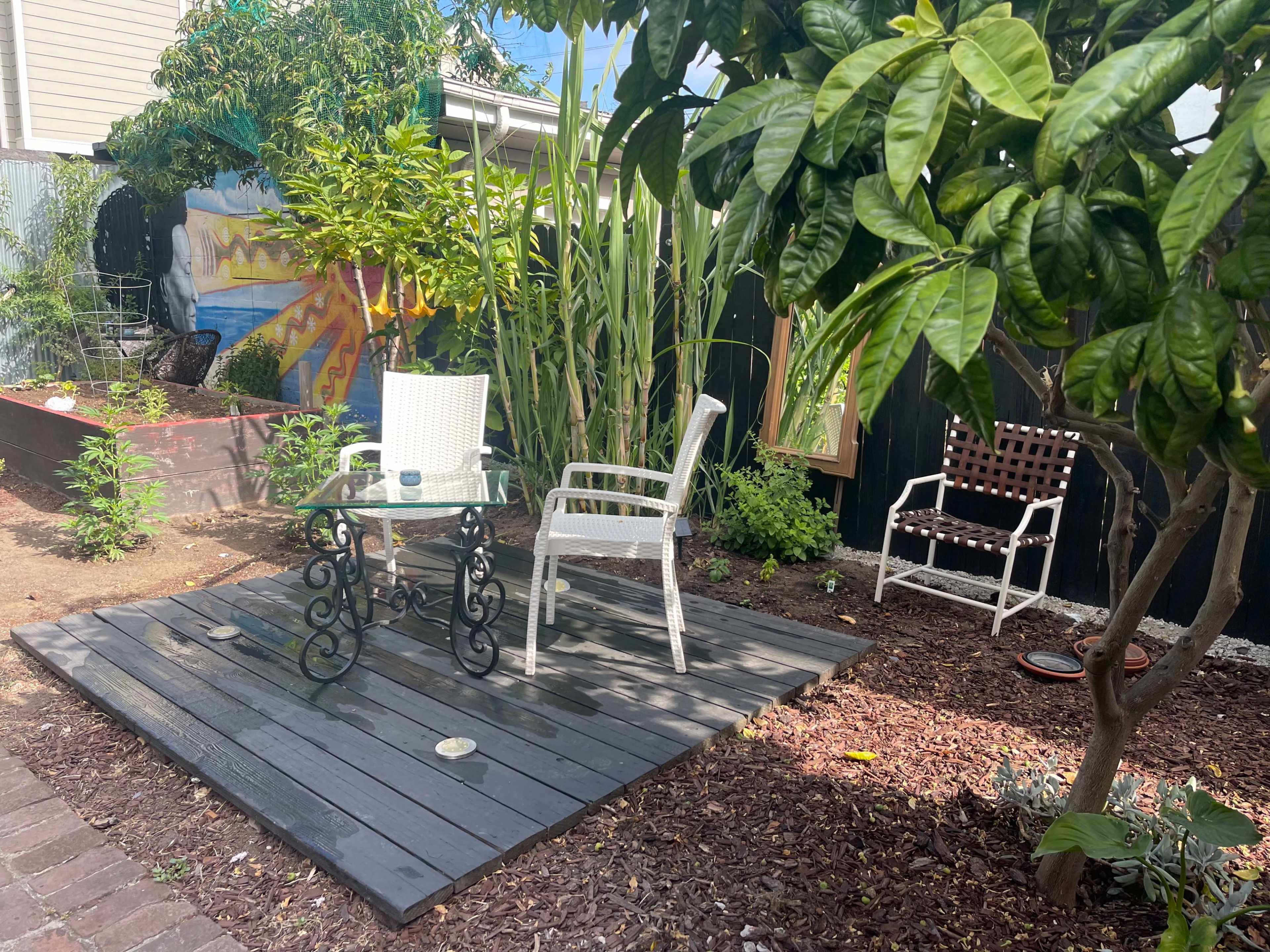 A small outdoor seating area features two chairs and a glass table on a wooden deck surrounded by plants and decorative artwork.
