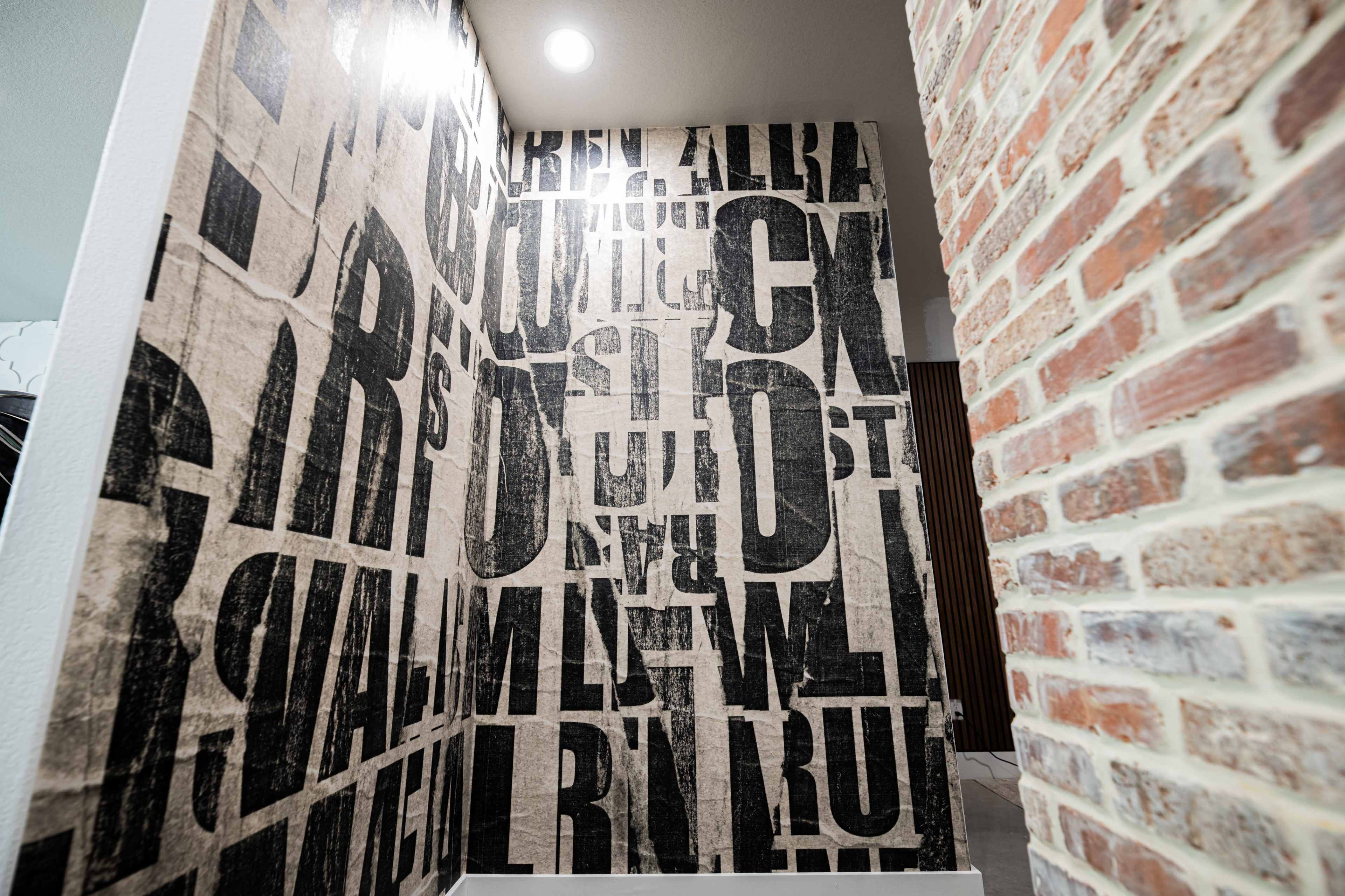 The image features a wall covered in large, bold black letters on a textured surface, adjacent to a section of exposed brick wall.