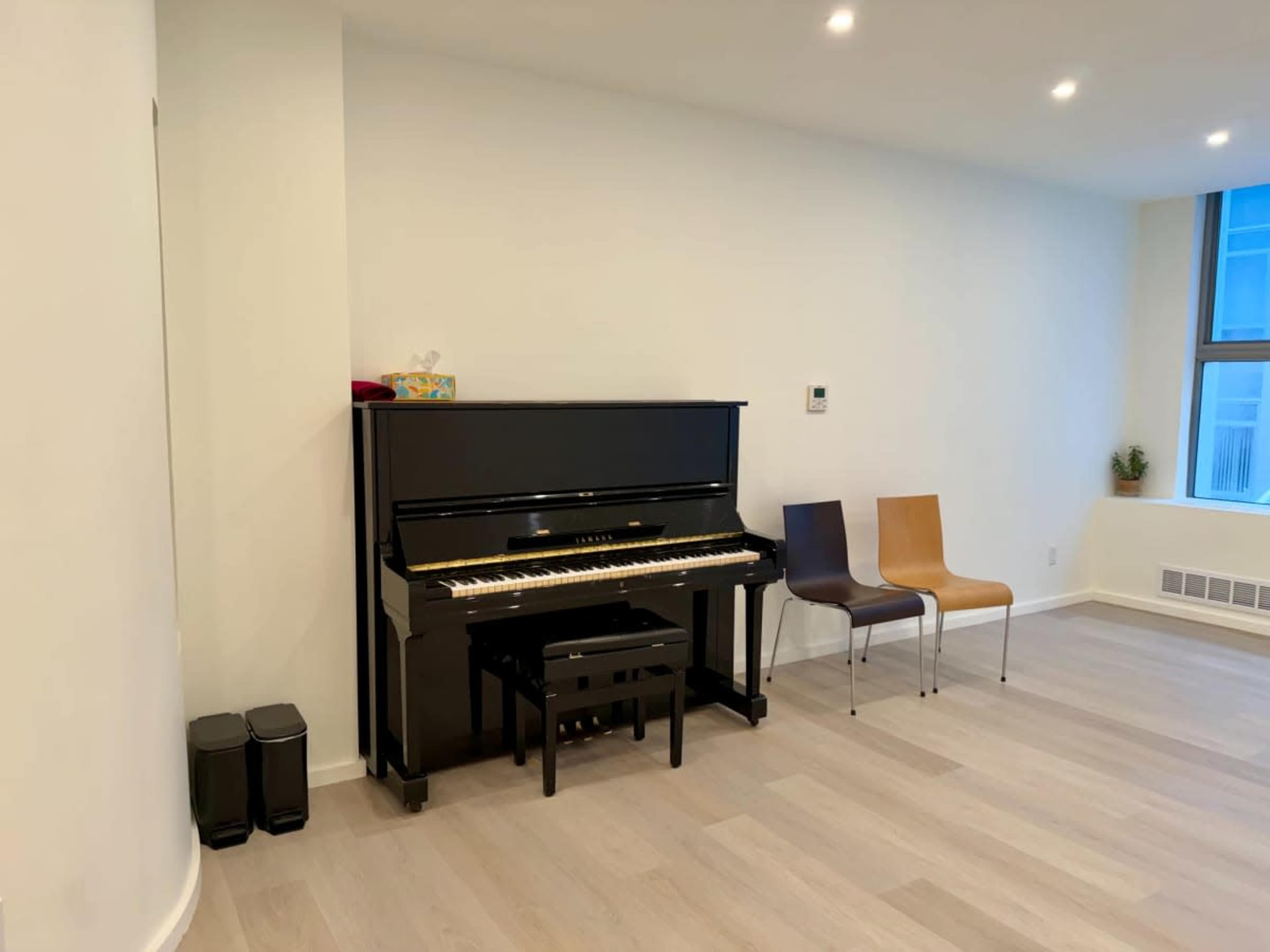 The image shows a black piano next to two wooden chairs in a sparsely furnished room with light-colored flooring.