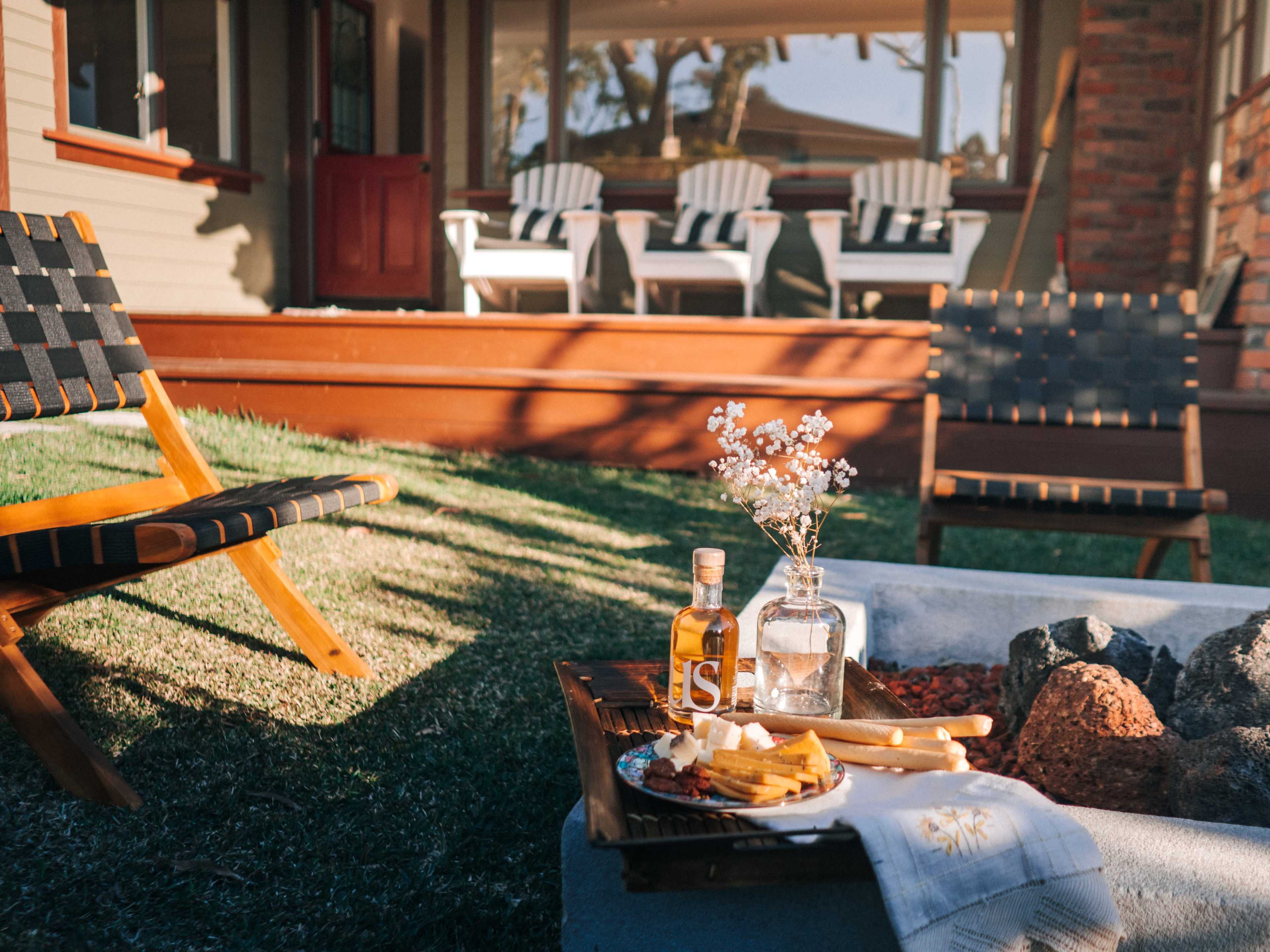 A small outdoor seating area features wooden chairs, a small table with a bottle and snacks, and a firepit surrounded by rocks.