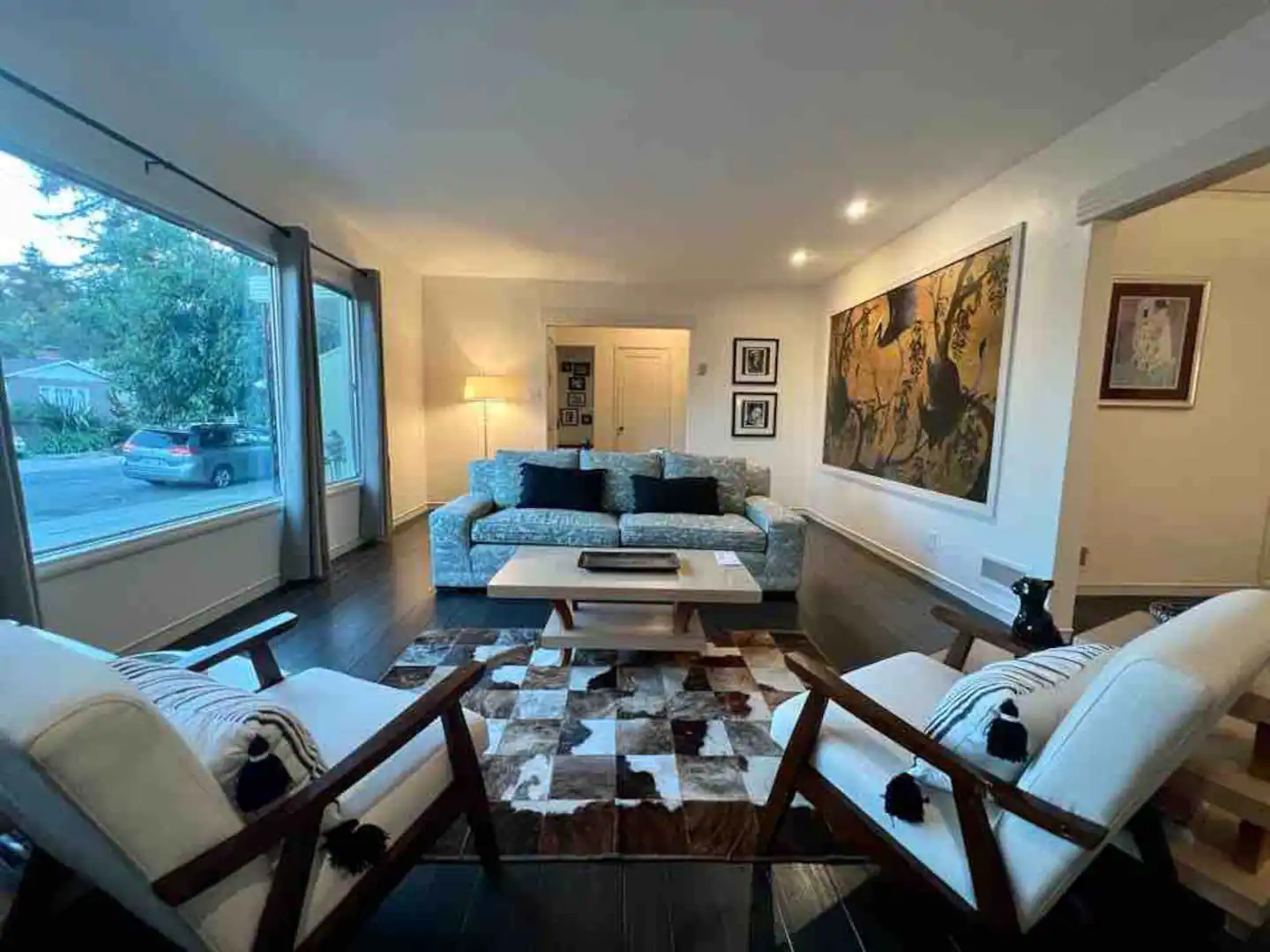 A cozy living room features a light blue sofa, two white chairs, a coffee table, and a large artwork on the wall, with a view through a large window.