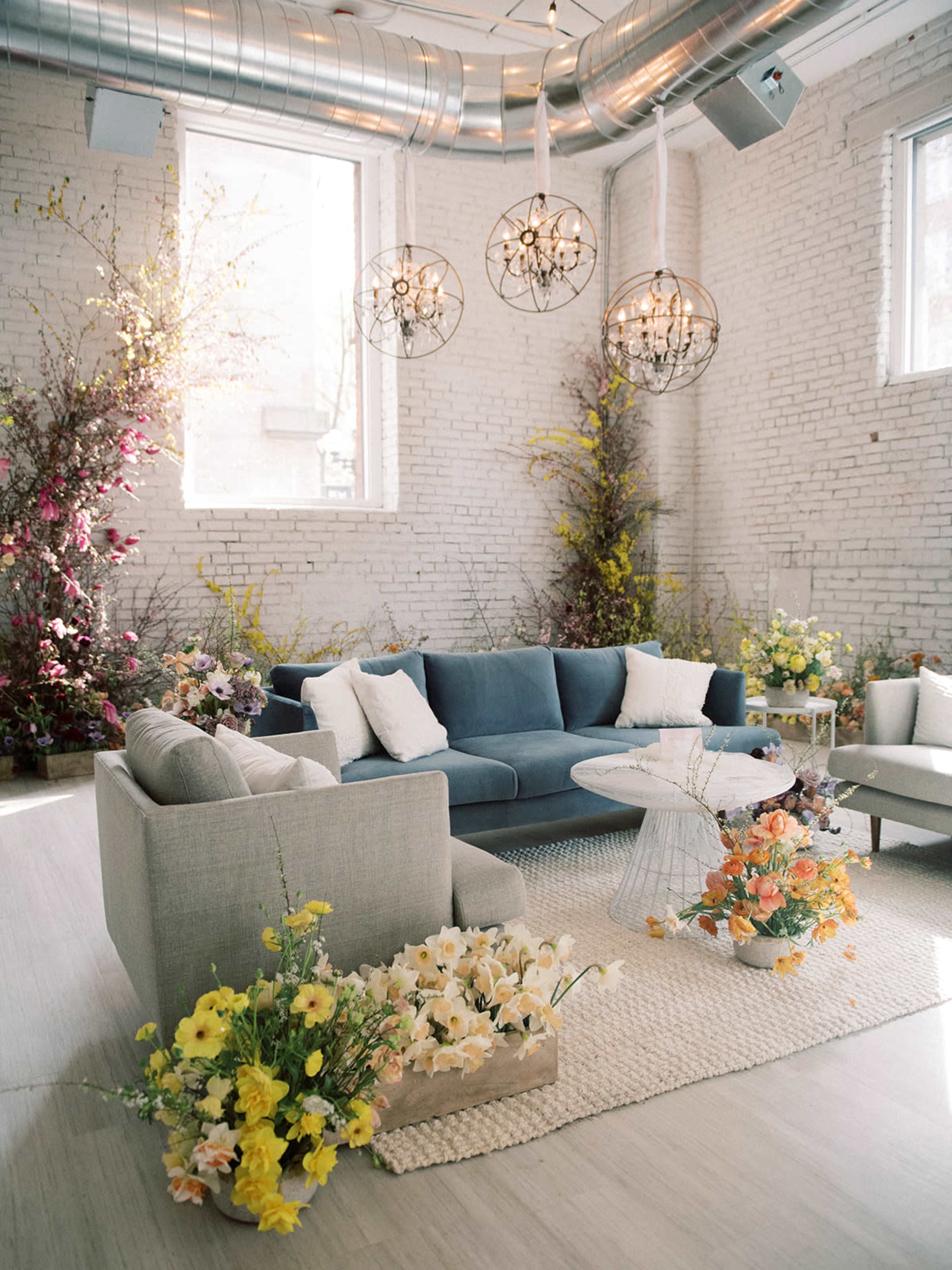 The image shows a bright, stylish indoor living space featuring a blue sectional sofa, a round coffee table, and various floral arrangements placed throughout the room.