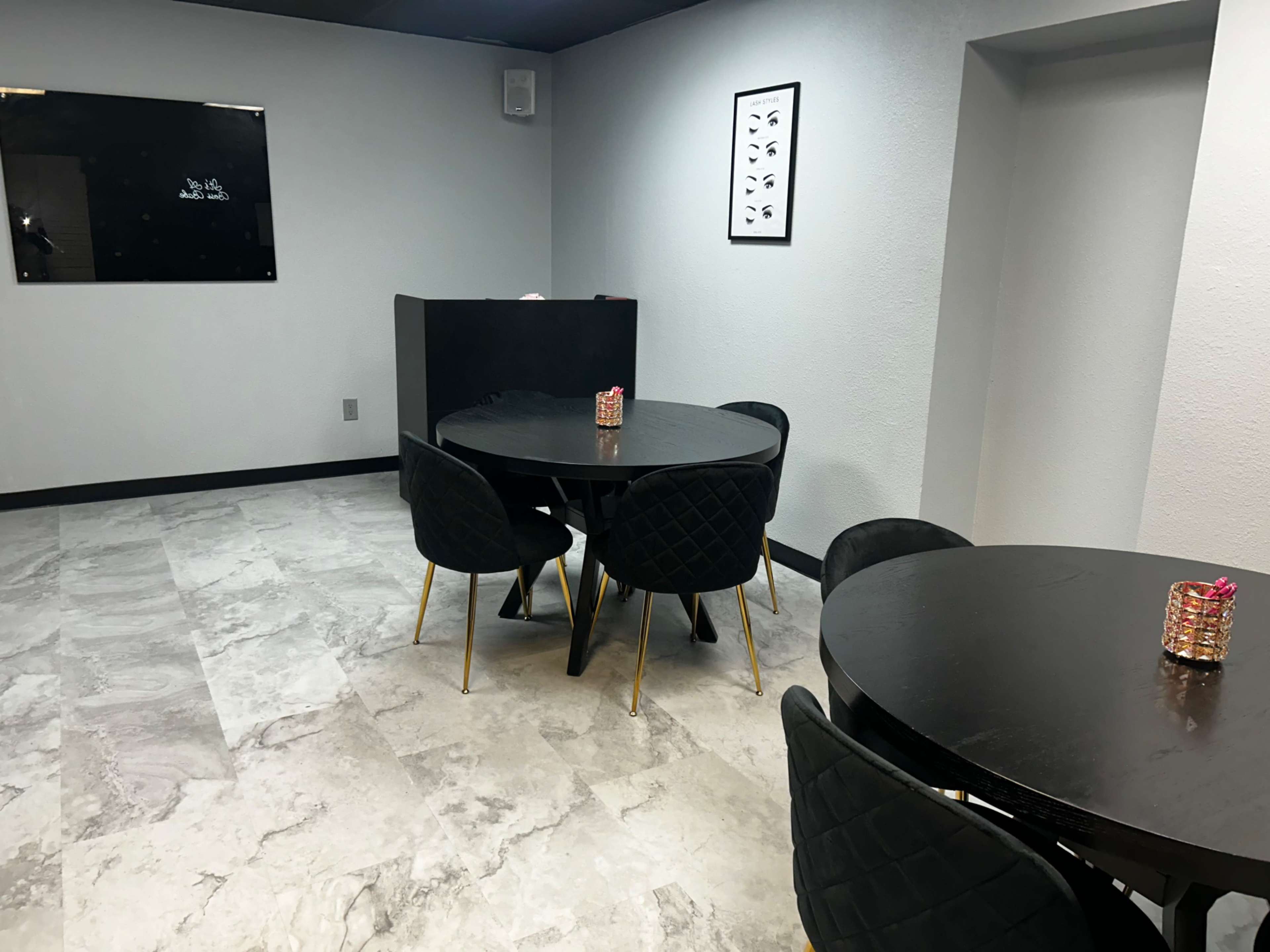 The image shows a modern, minimalist room featuring two round black tables surrounded by black chairs, with gray tile flooring and light walls.