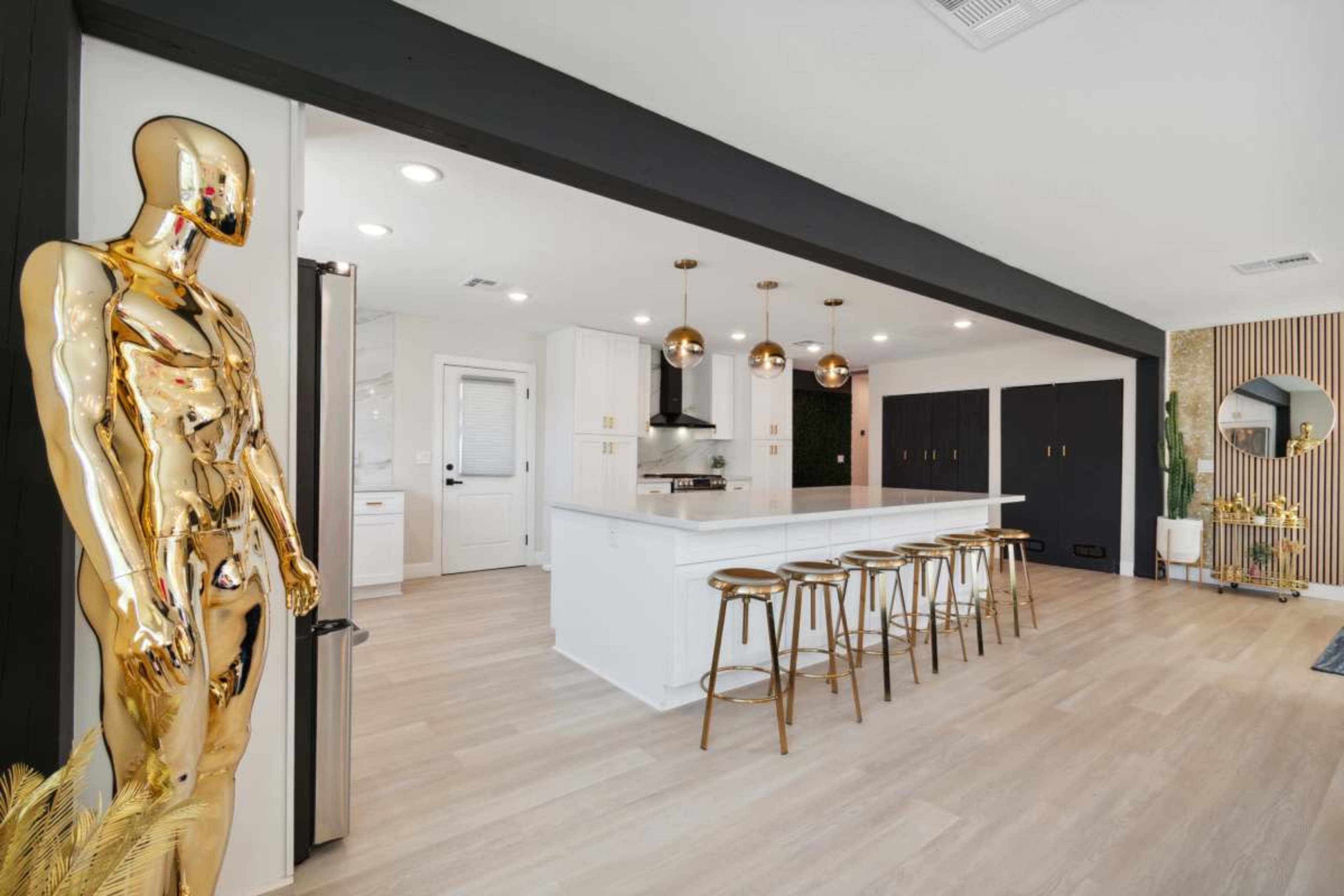 A modern kitchen features white cabinetry, a large island with gold bar stools, and a golden mannequin standing nearby.