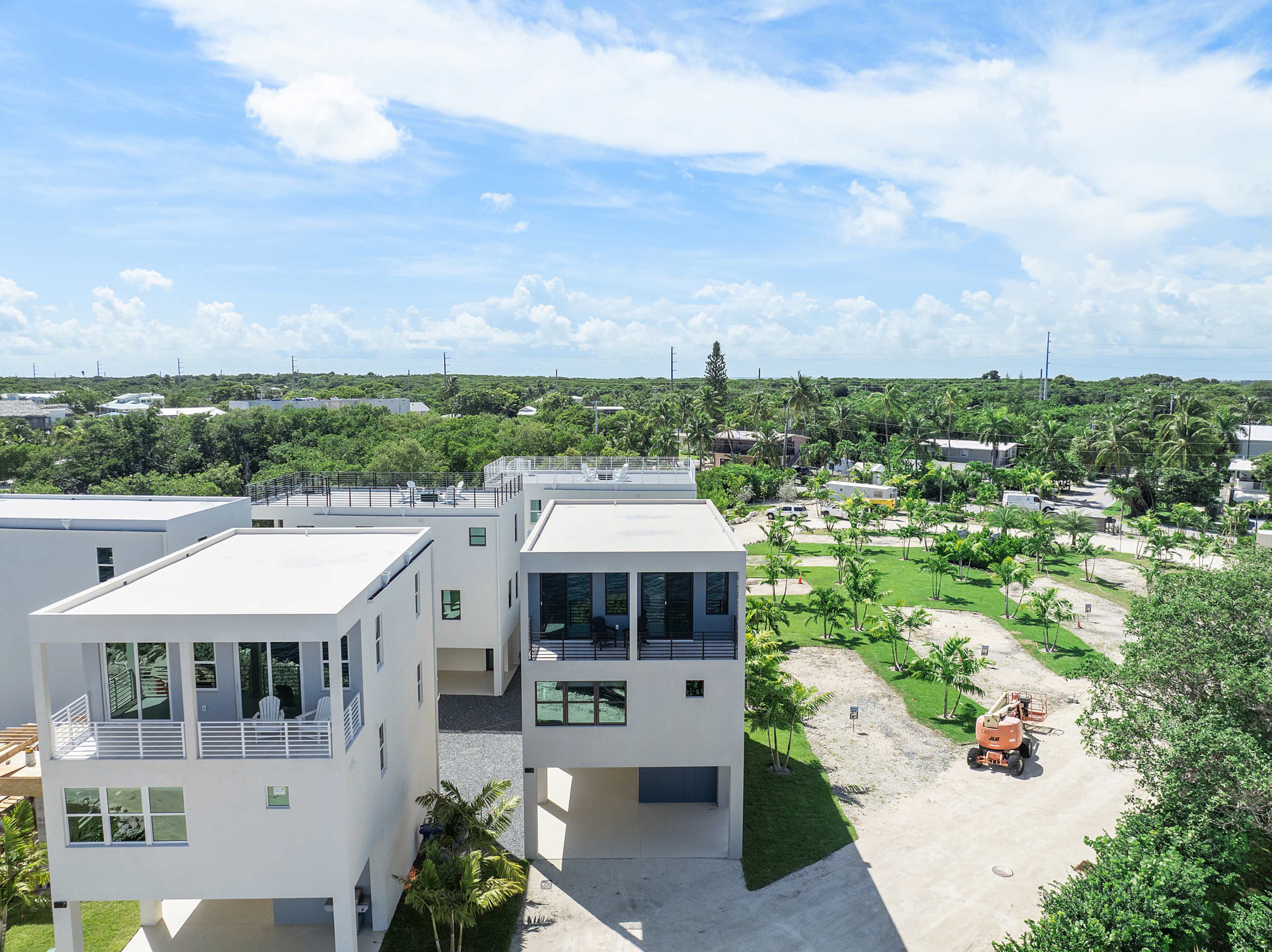 The image shows a construction site with modern houses surrounded by greenery and a clear sky.