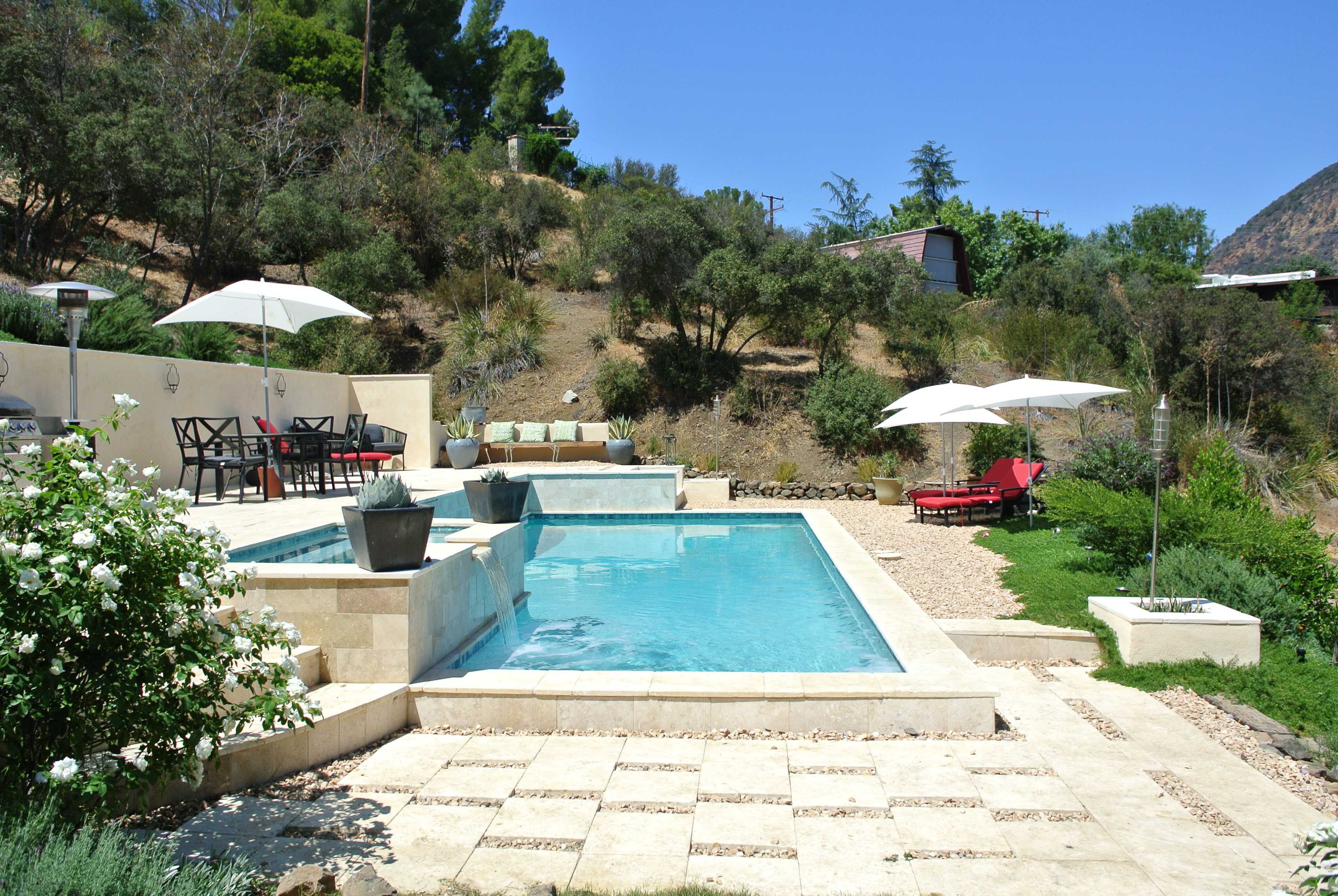A landscaped outdoor area featuring a swimming pool, lounge chairs, and patio seating under umbrellas, surrounded by natural greenery and a hillside.