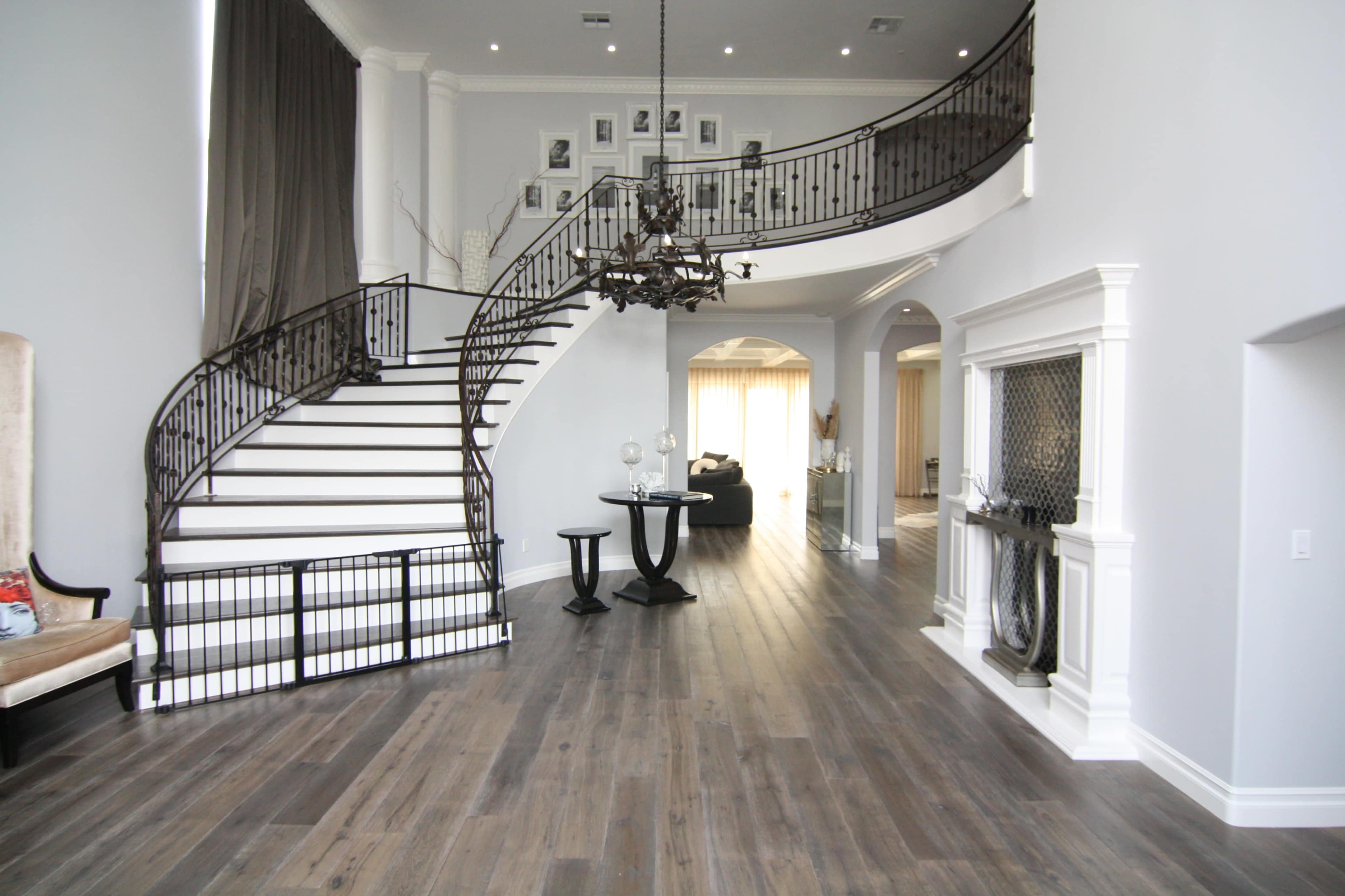 The image shows a spacious, modern foyer featuring a curved staircase with a wrought-iron railing, a chandelier, and a seating area.