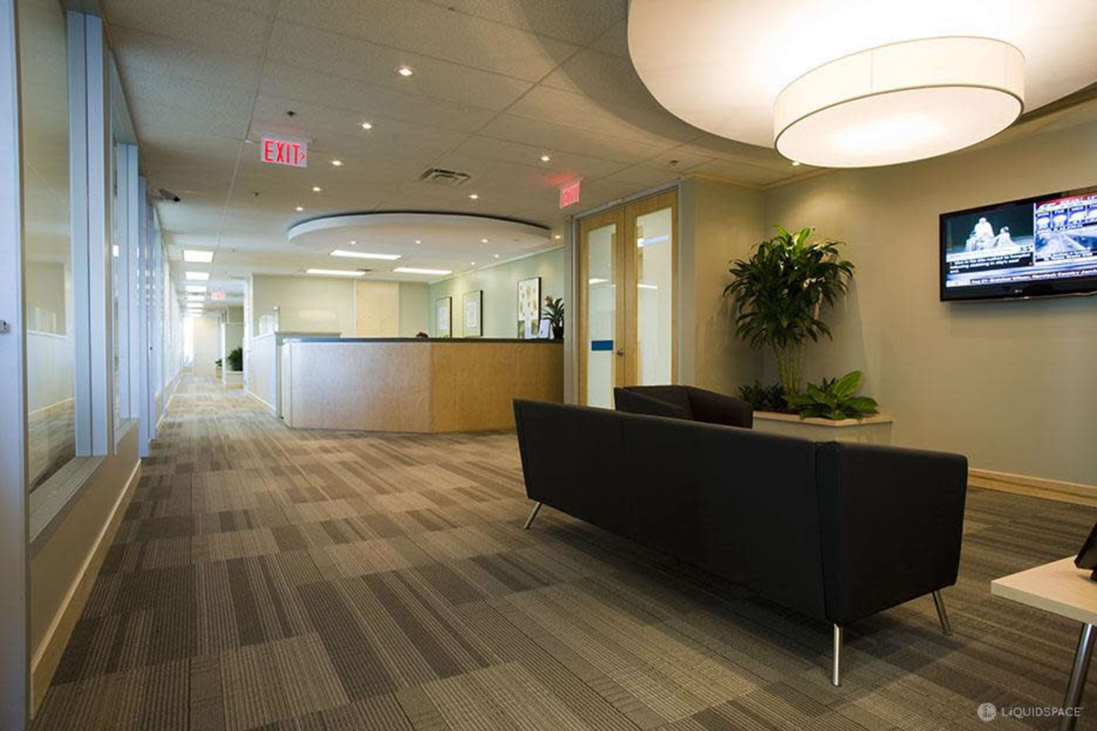 A modern office reception area with a long hallway, a couch, a large circular light fixture, and a television displaying news.