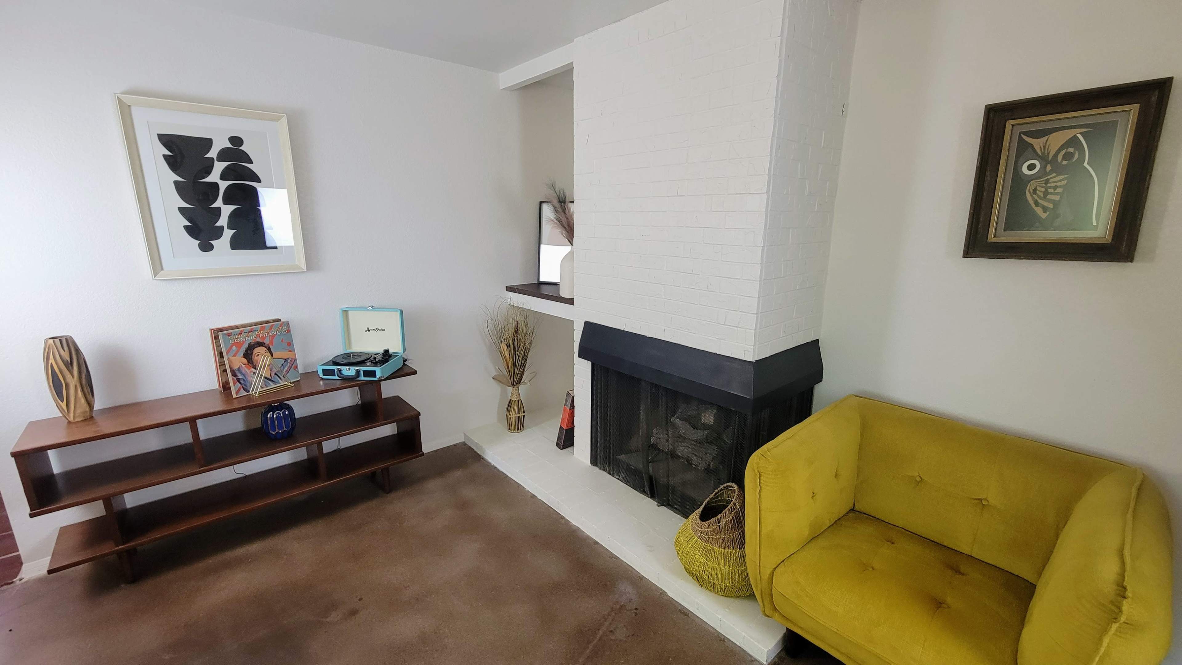 A living room with a yellow couch, a modern art piece hanging on the wall, a white fireplace, and a wooden shelf displaying decorative items.