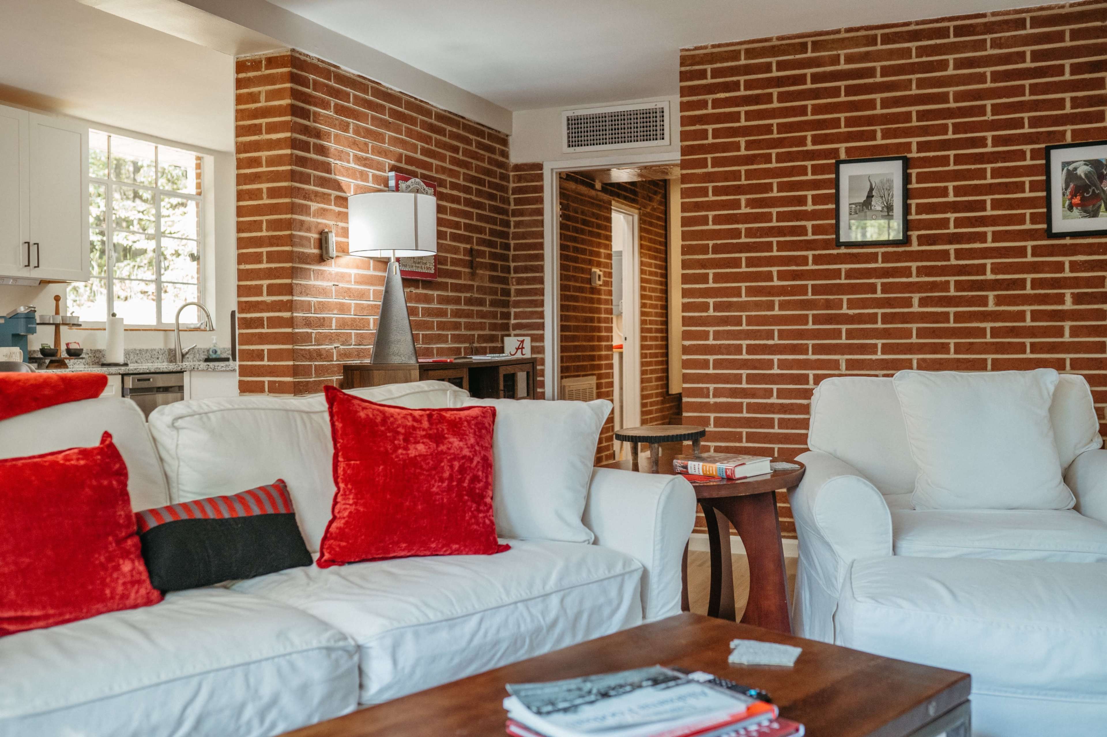 The living area features white couches with red accent pillows, a wooden coffee table, and exposed brick walls, creating a cozy atmosphere.