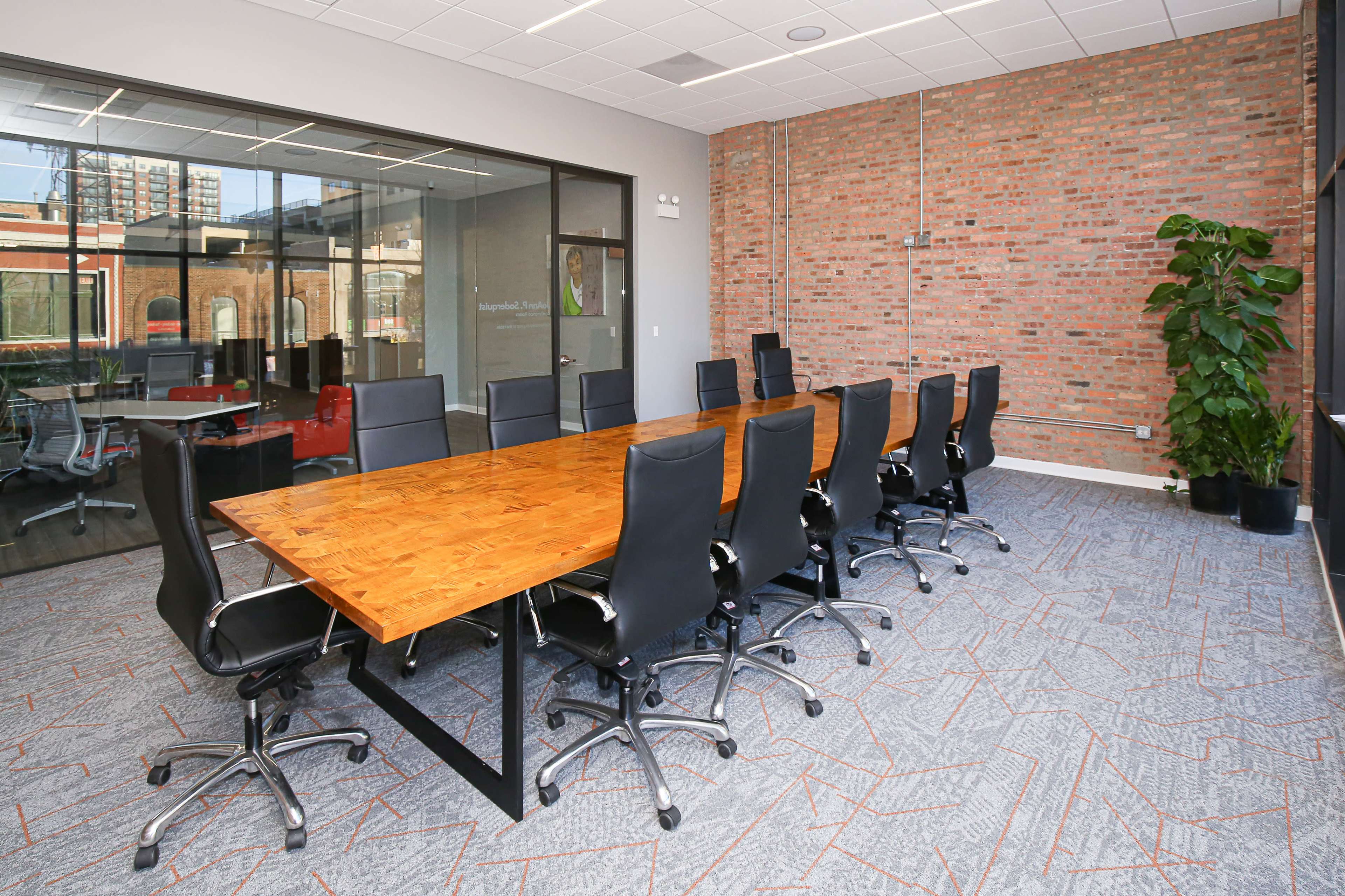 A spacious conference room featuring a long wooden table surrounded by black ergonomic chairs, with a large glass window and a plant in the corner.