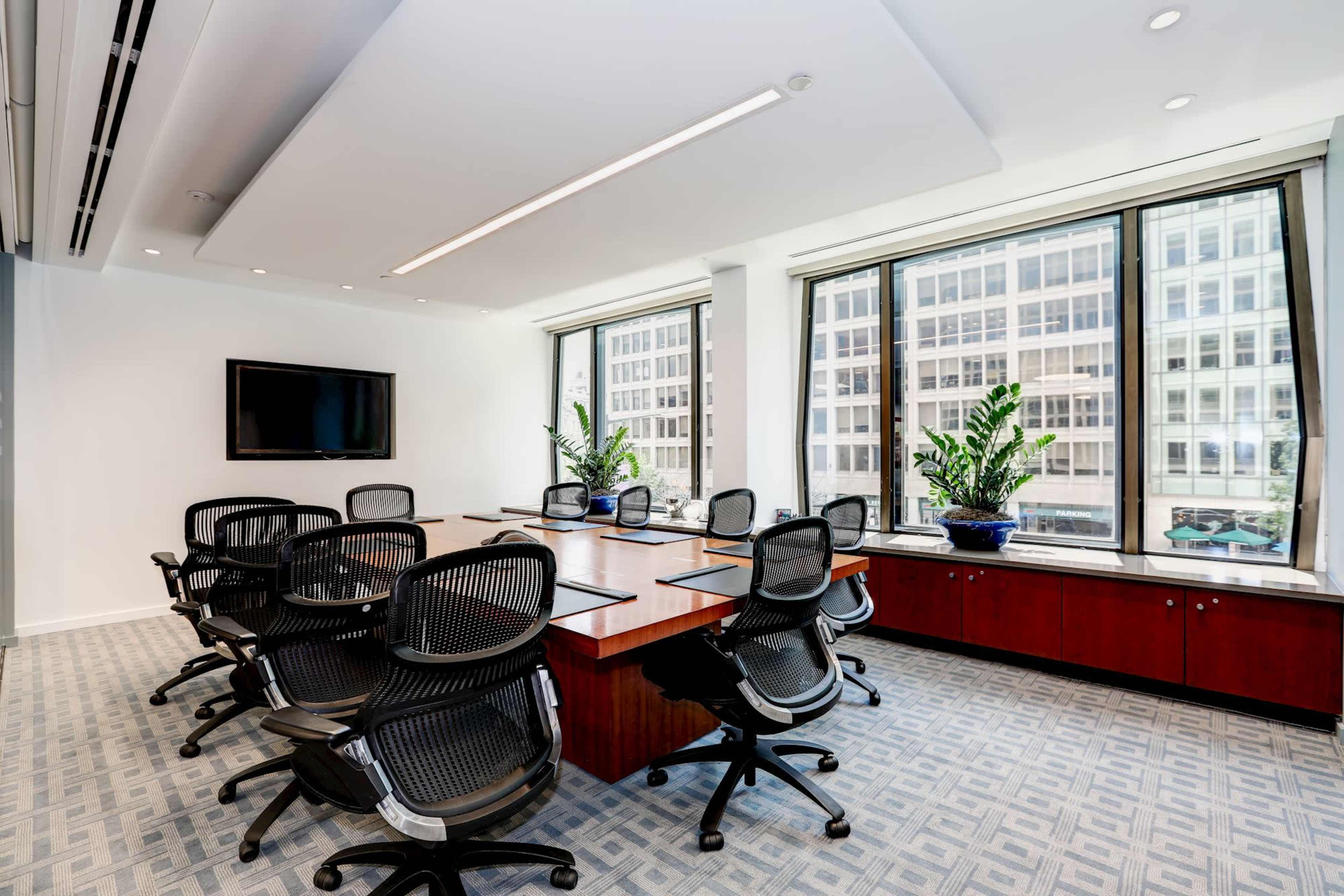 A modern conference room with a large wooden table surrounded by office chairs and three windows overlooking a cityscape.