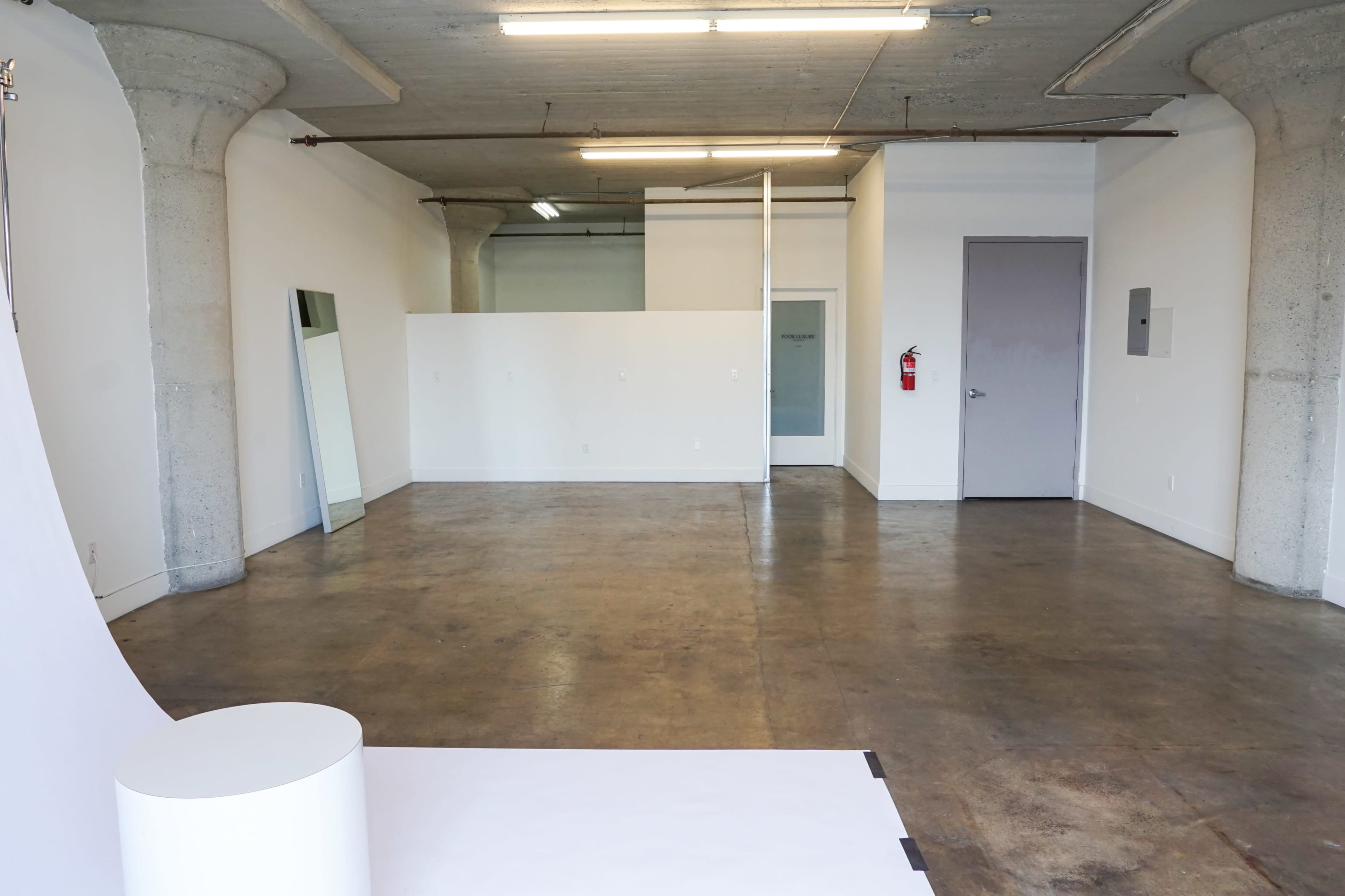 The image shows an empty room with concrete pillars, polished concrete flooring, and white walls, featuring a dividing wall and a mirror on one side.