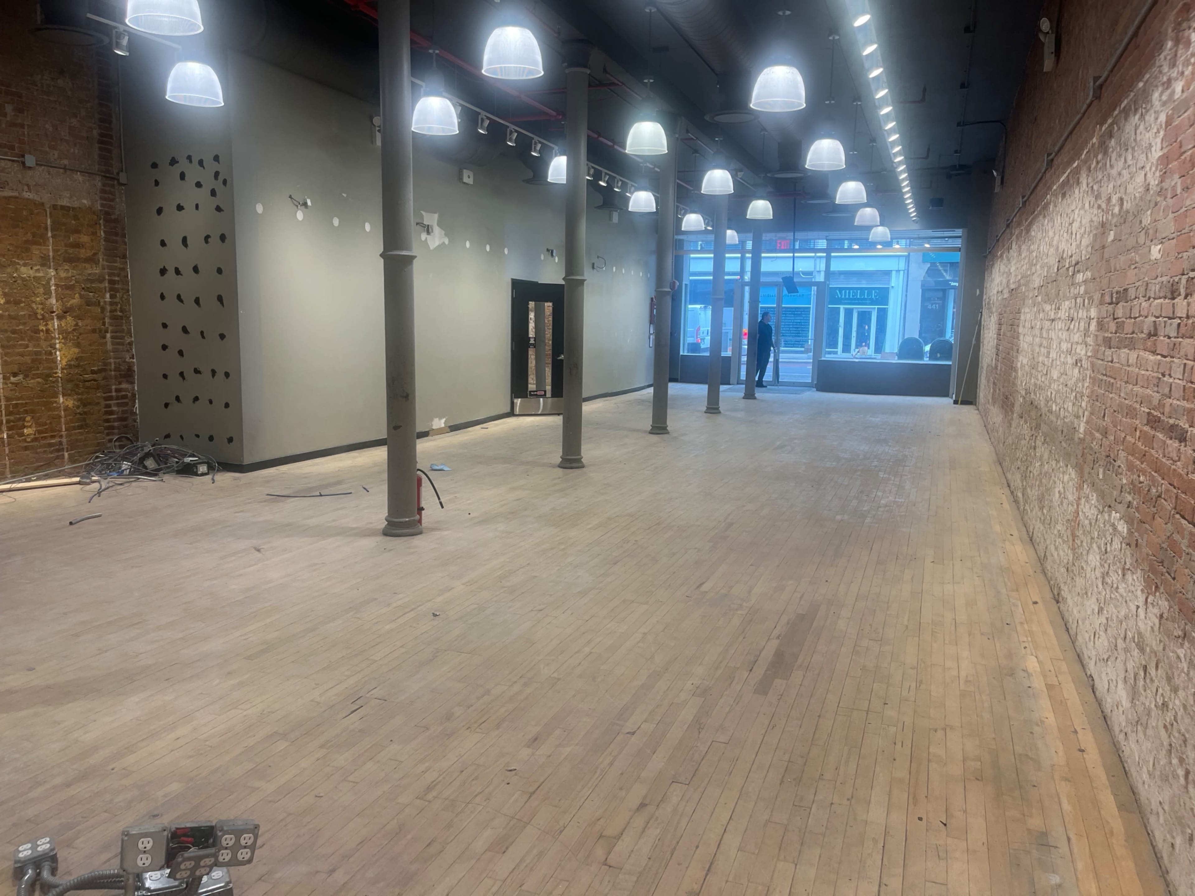The image shows an empty commercial space with wooden floors, brick walls, and overhead lights, along with a person standing at the far end.