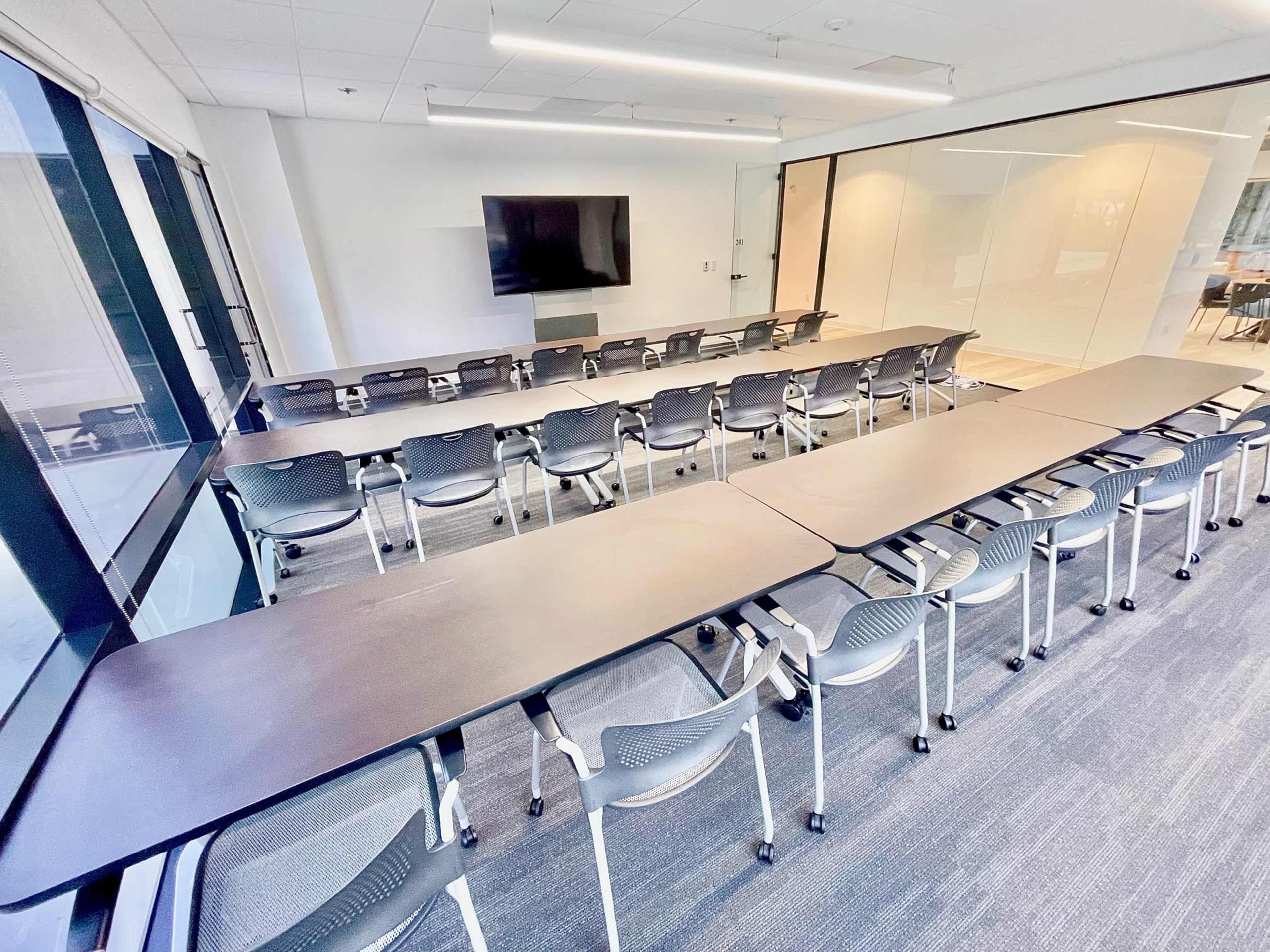 A conference room features a long arrangement of rectangular tables with chairs, and a large screen on the wall.