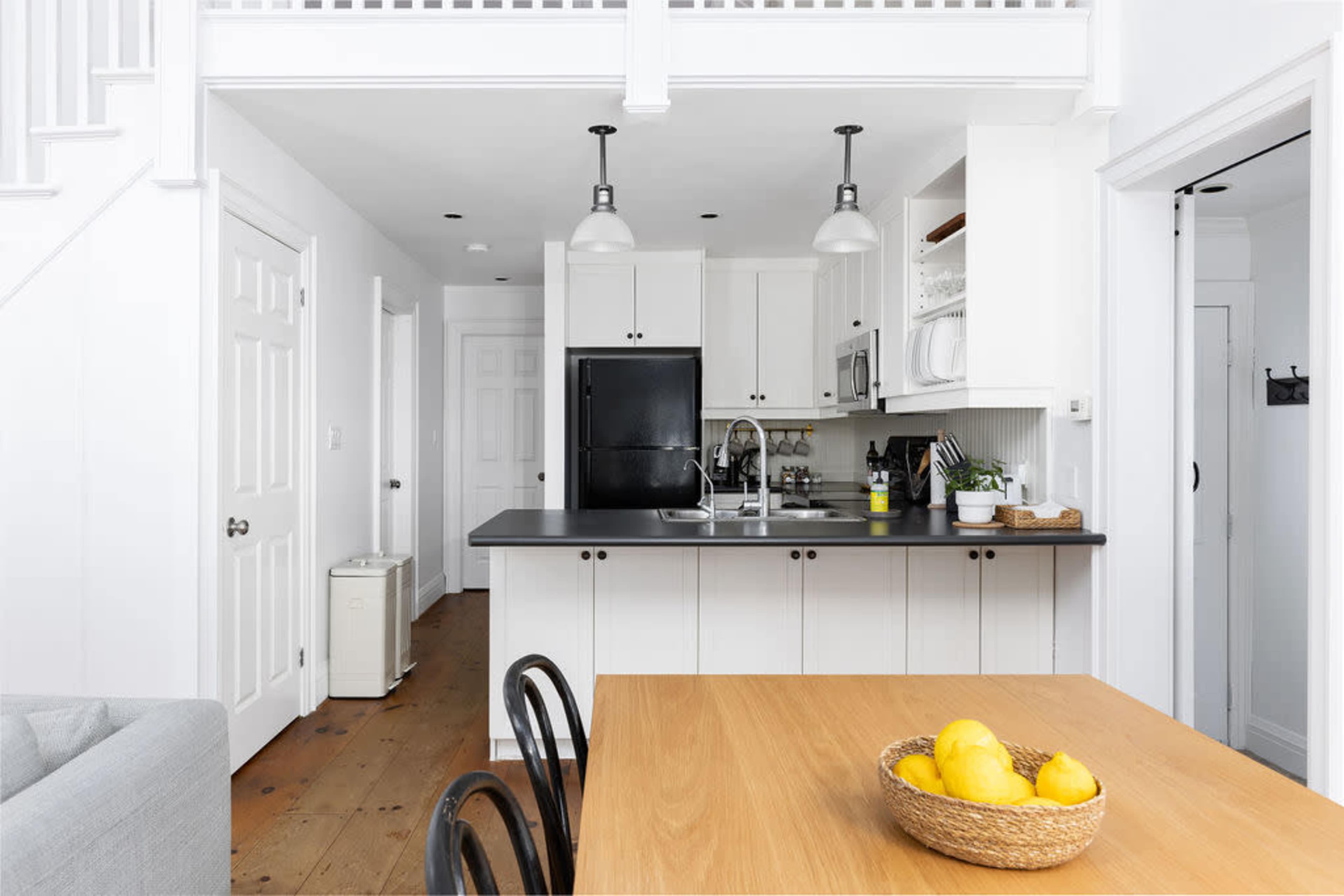 The image shows a modern kitchen with a small dining area featuring a wooden table, black chairs, and a bowl of lemons.