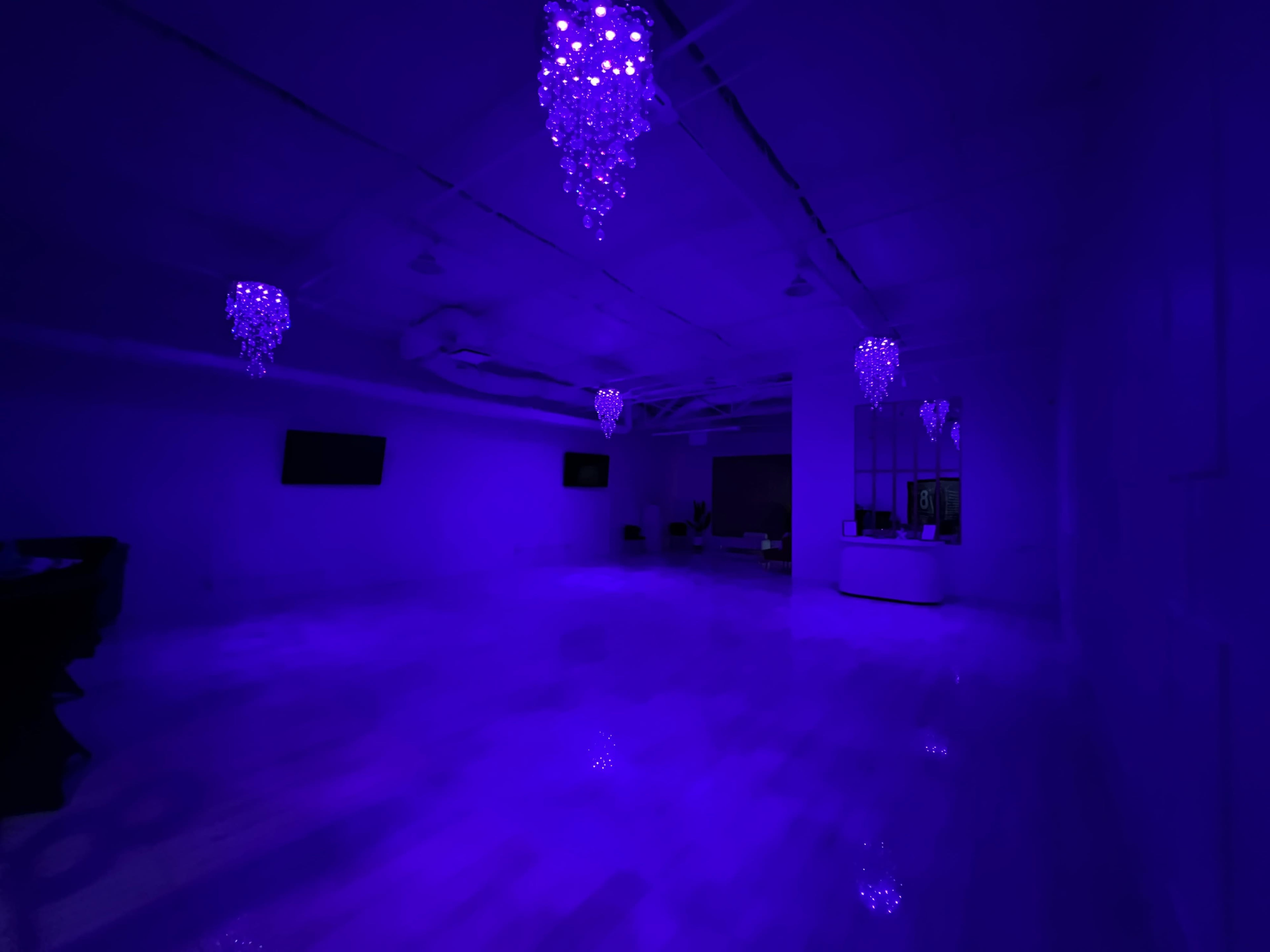 A large, empty room is illuminated in purple light, featuring several chandelier-like fixtures on the ceiling.