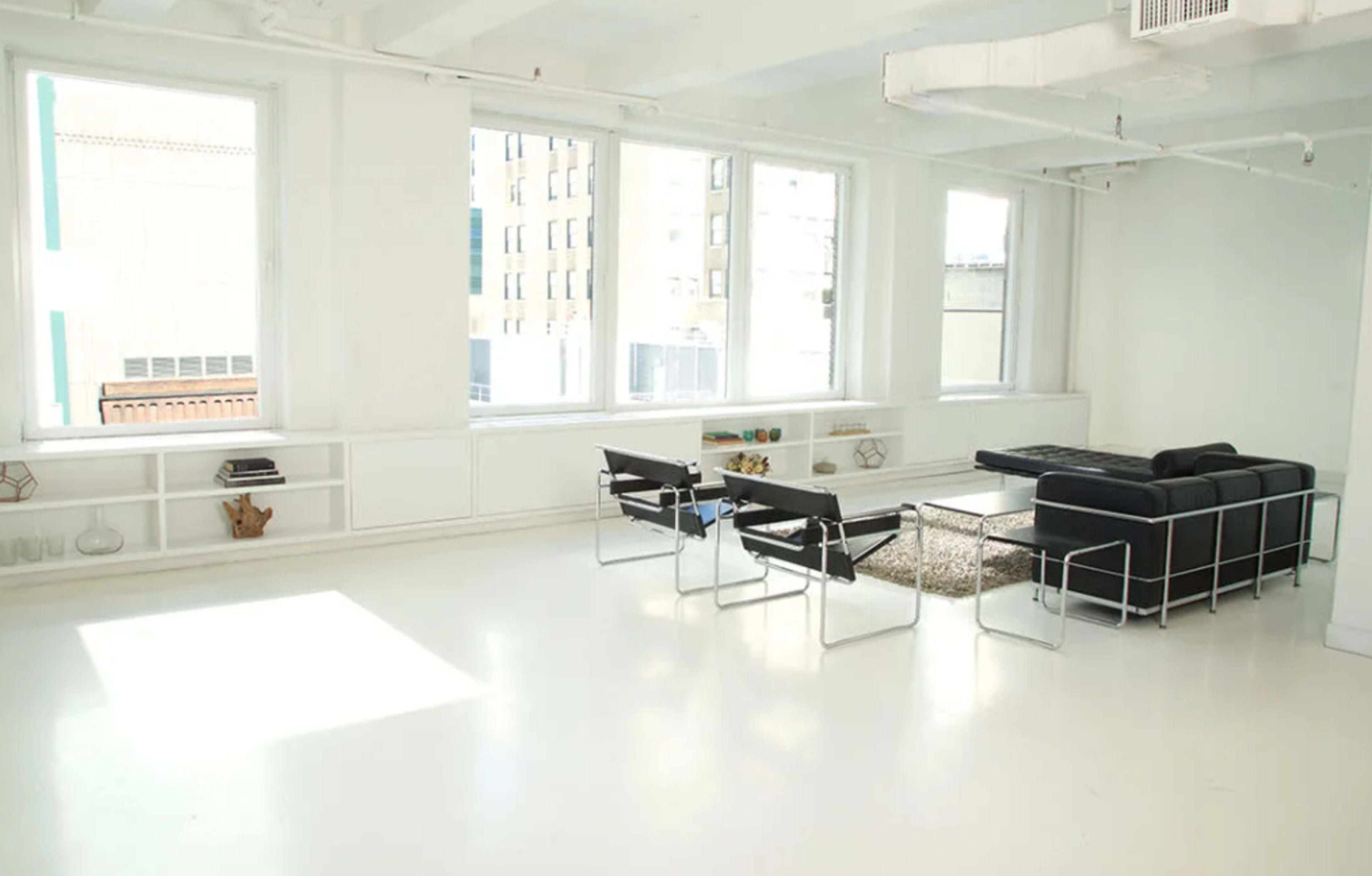 A minimalist living space features a black sofa, two chairs, a coffee table, and large windows providing natural light.