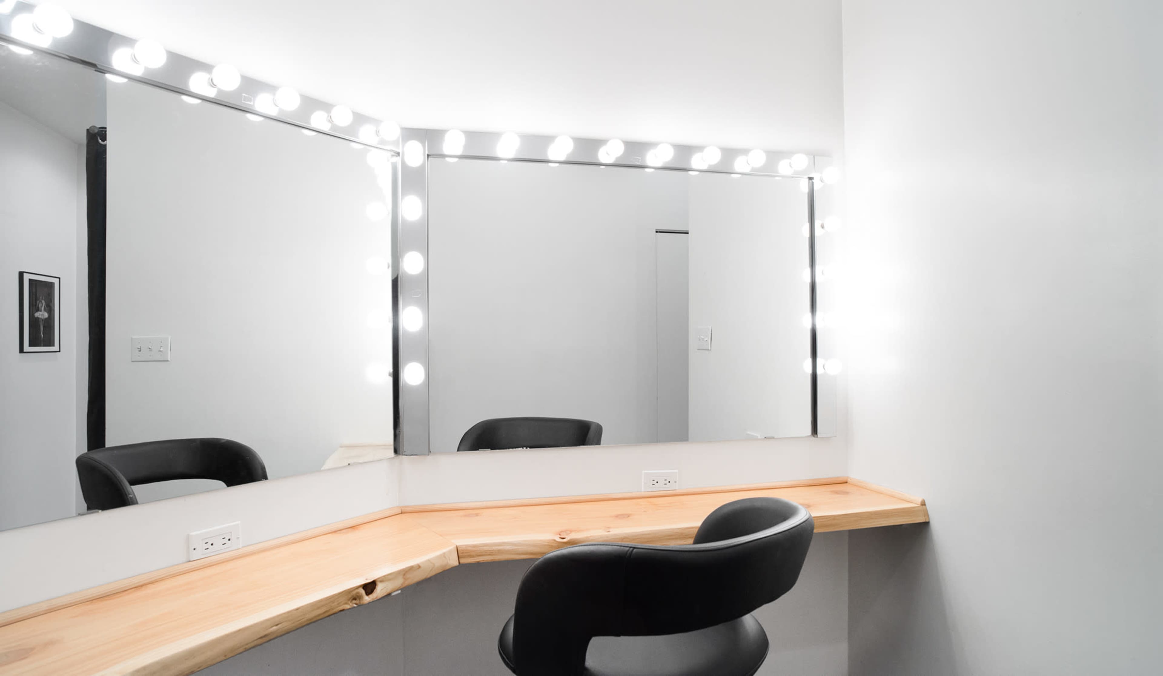 A makeup vanity area features a wooden countertop and three mirrors surrounded by bright lights.