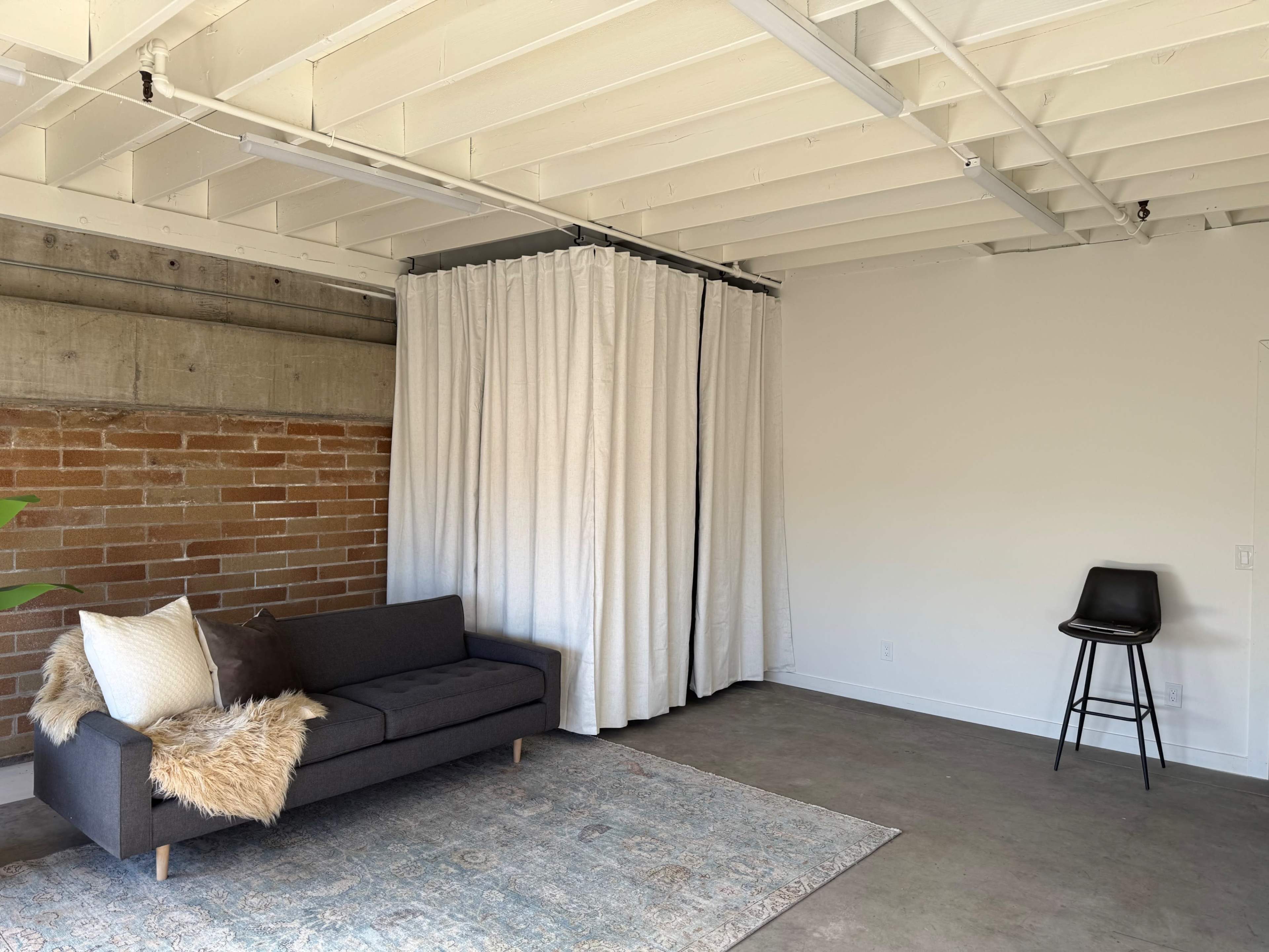 The image shows a minimalist living space featuring a dark sofa, a large curtain, a single black stool, and a textured area rug on a concrete floor.