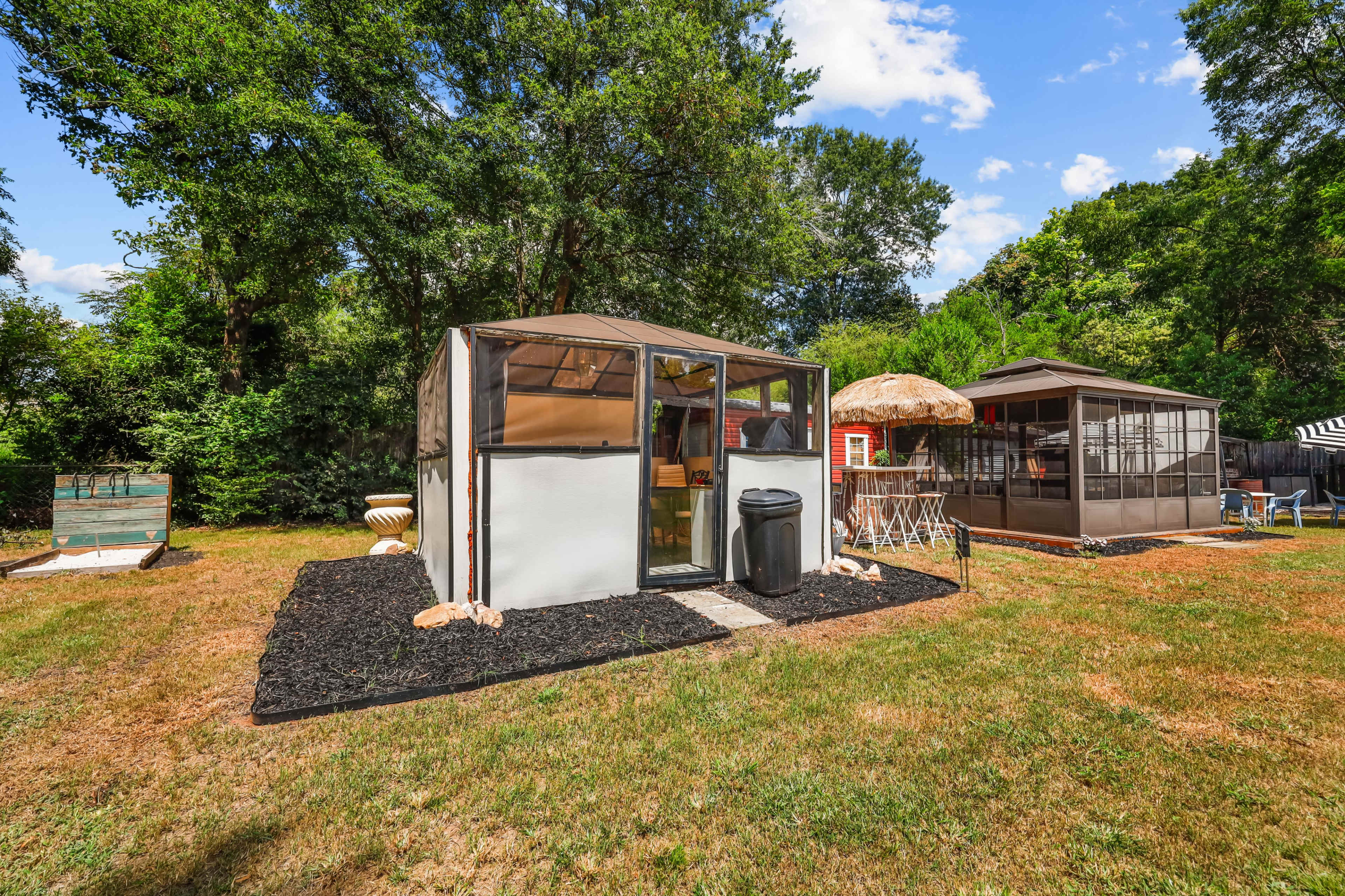 The image shows two screened structures in a yard with grassy areas, surrounded by trees under a blue sky.