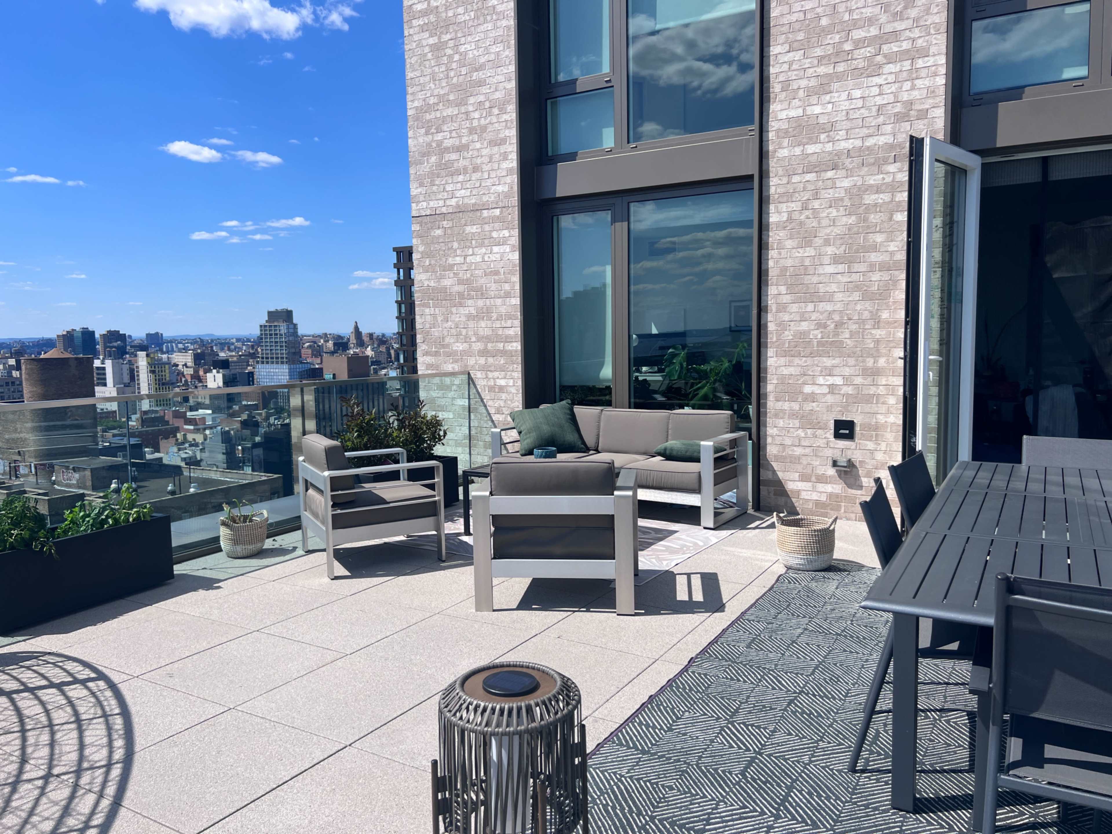 A modern rooftop terrace featuring gray furniture, a dining table, and city views under a clear blue sky.