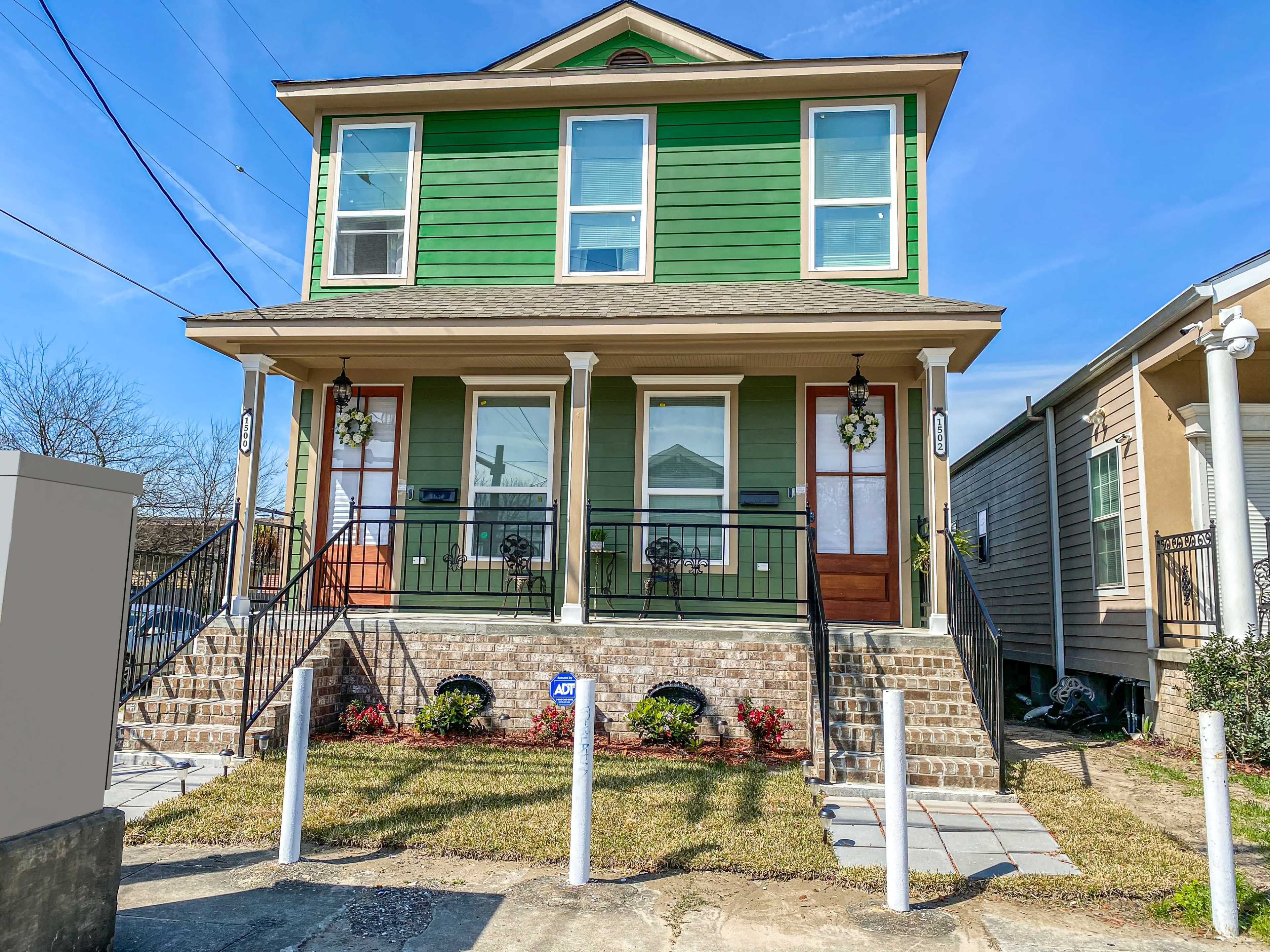A two-story green duplex with a brick foundation features two entrance doors, each with a small porch and decorative plants.