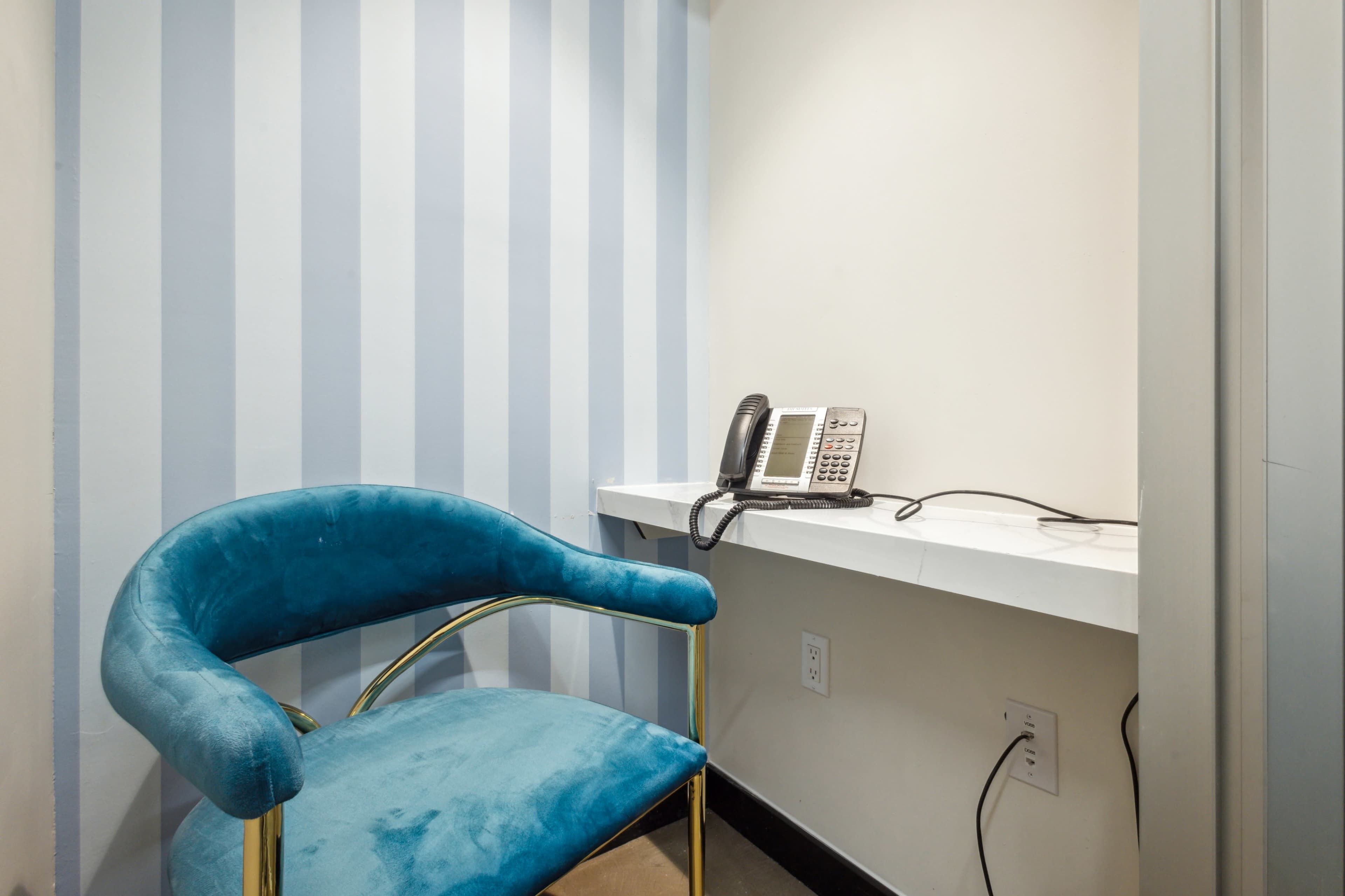 A blue upholstered chair is positioned next to a wall-mounted telephone on a white desk in a room with striped walls.