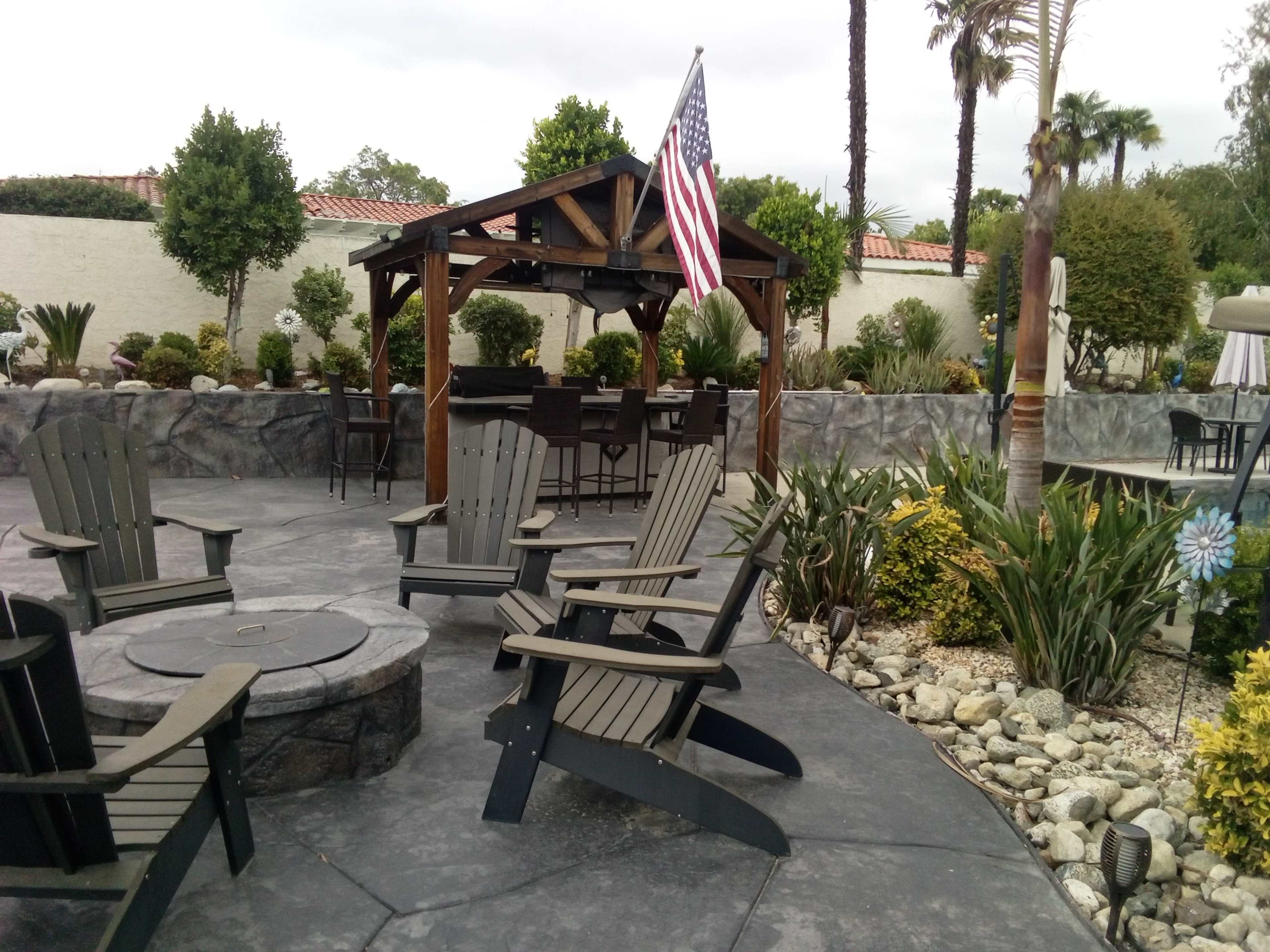 The image shows a patio area with a wooden gazebo featuring an American flag, surrounded by several potted plants and seating arranged around a fire pit.