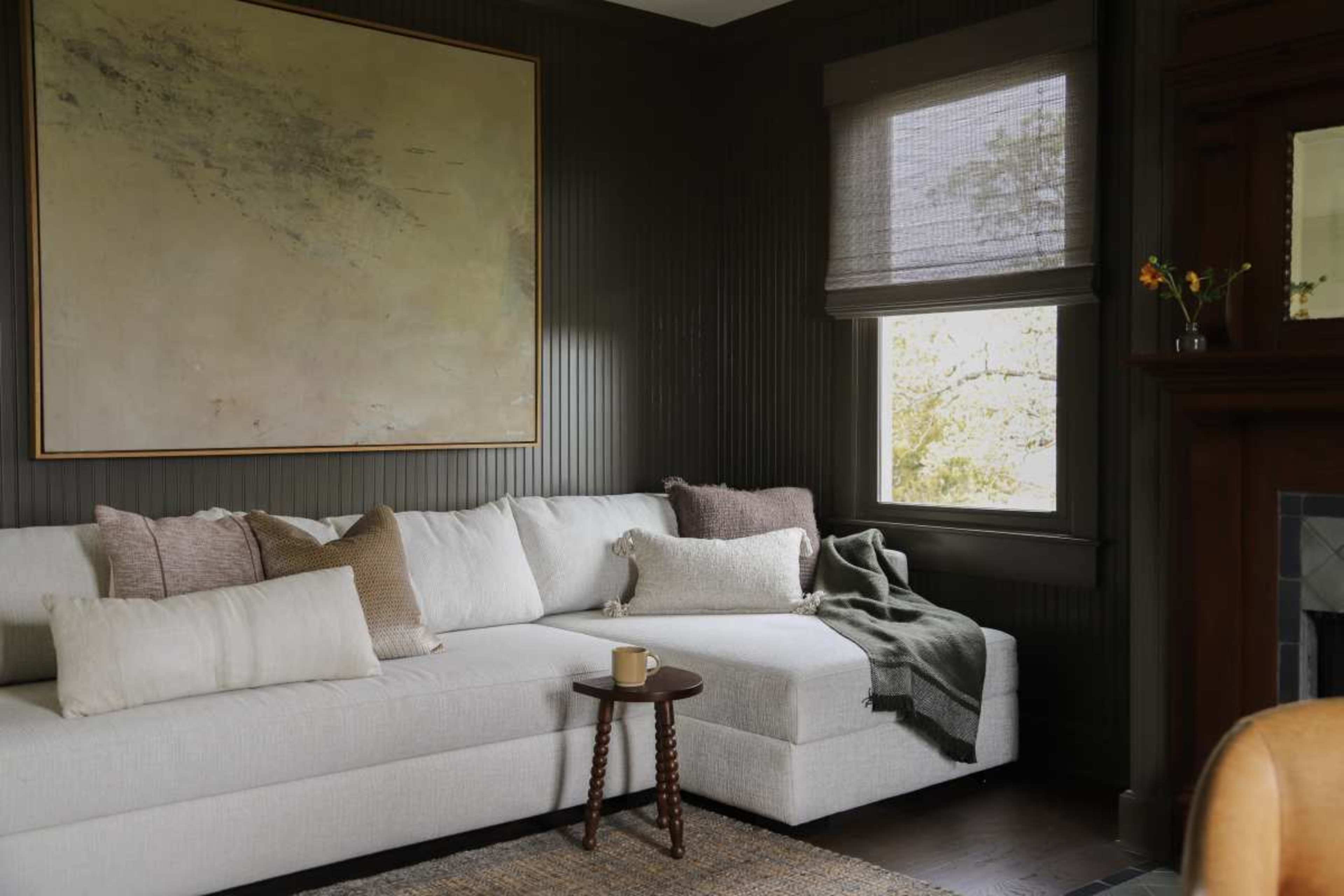The image shows a light-colored sectional sofa with various textured pillows, a small wooden side table, and a window with shades, all set against dark paneled walls.