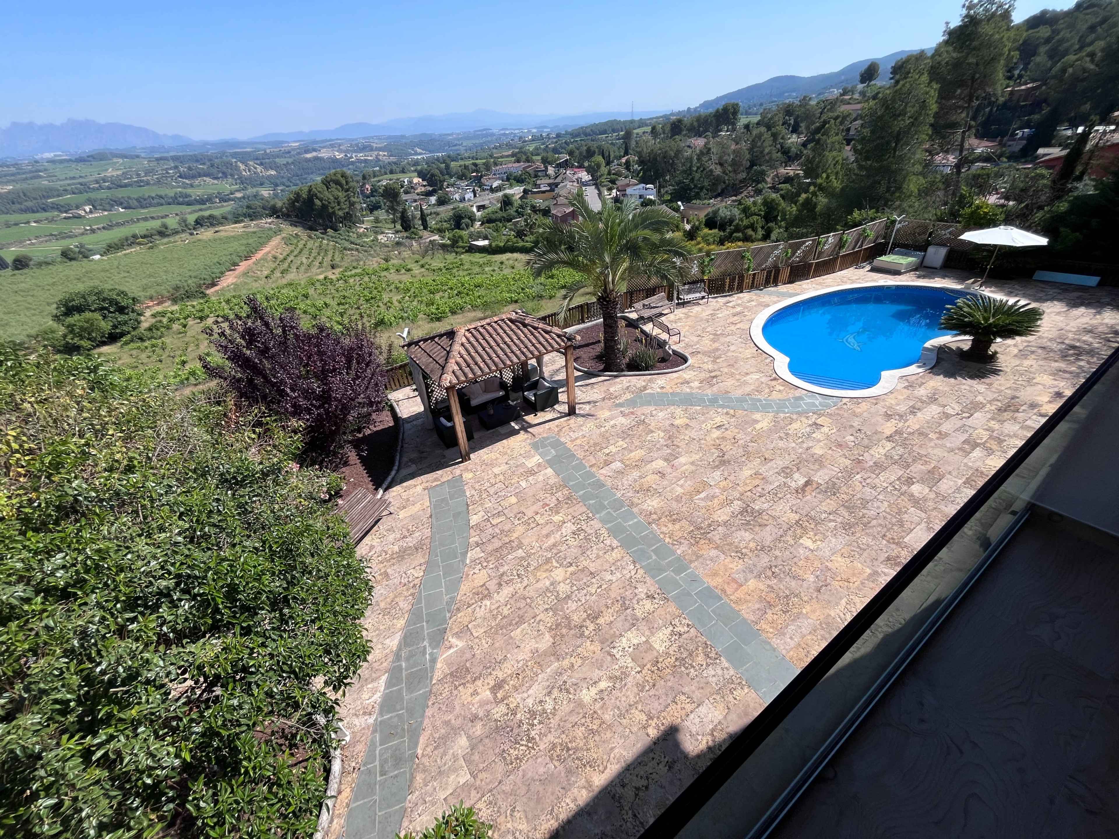 The image shows a landscaped backyard with a swimming pool, a gazebo, and a scenic view of rolling hills and a distant village.