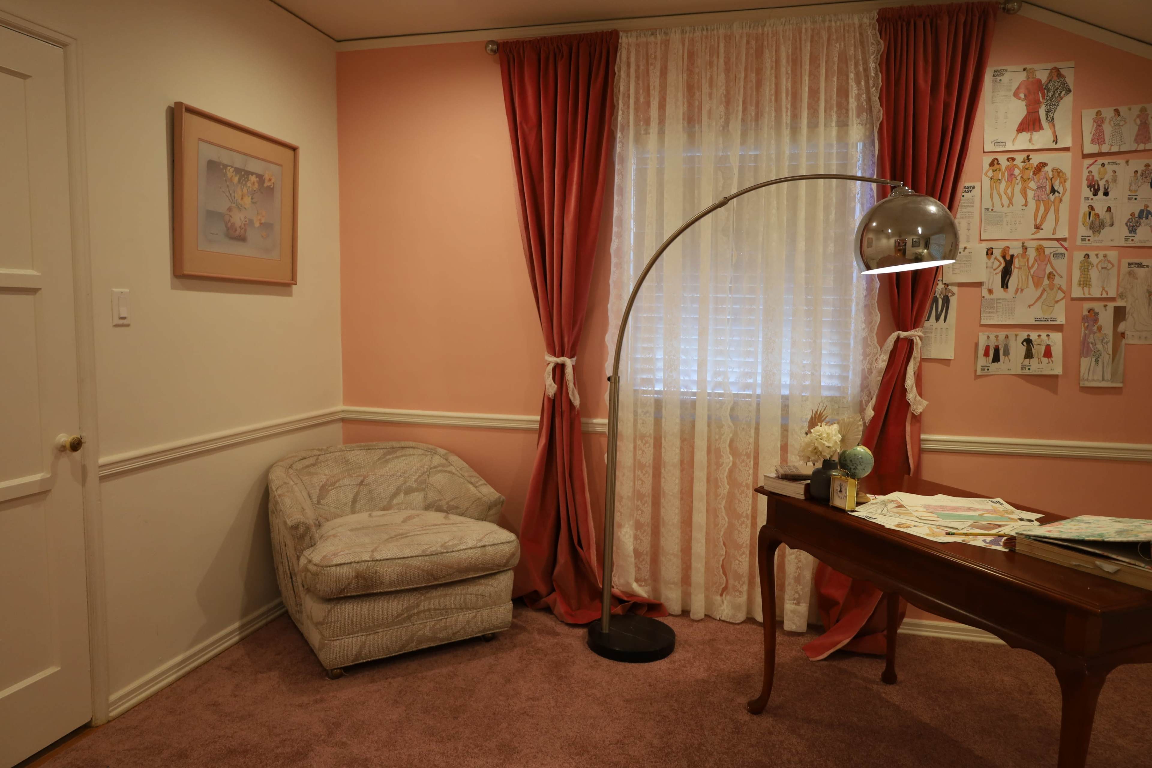 The image shows a softly lit room with pink walls, a patterned armchair, a floor lamp, a desk covered with papers, and curtains draping a window.