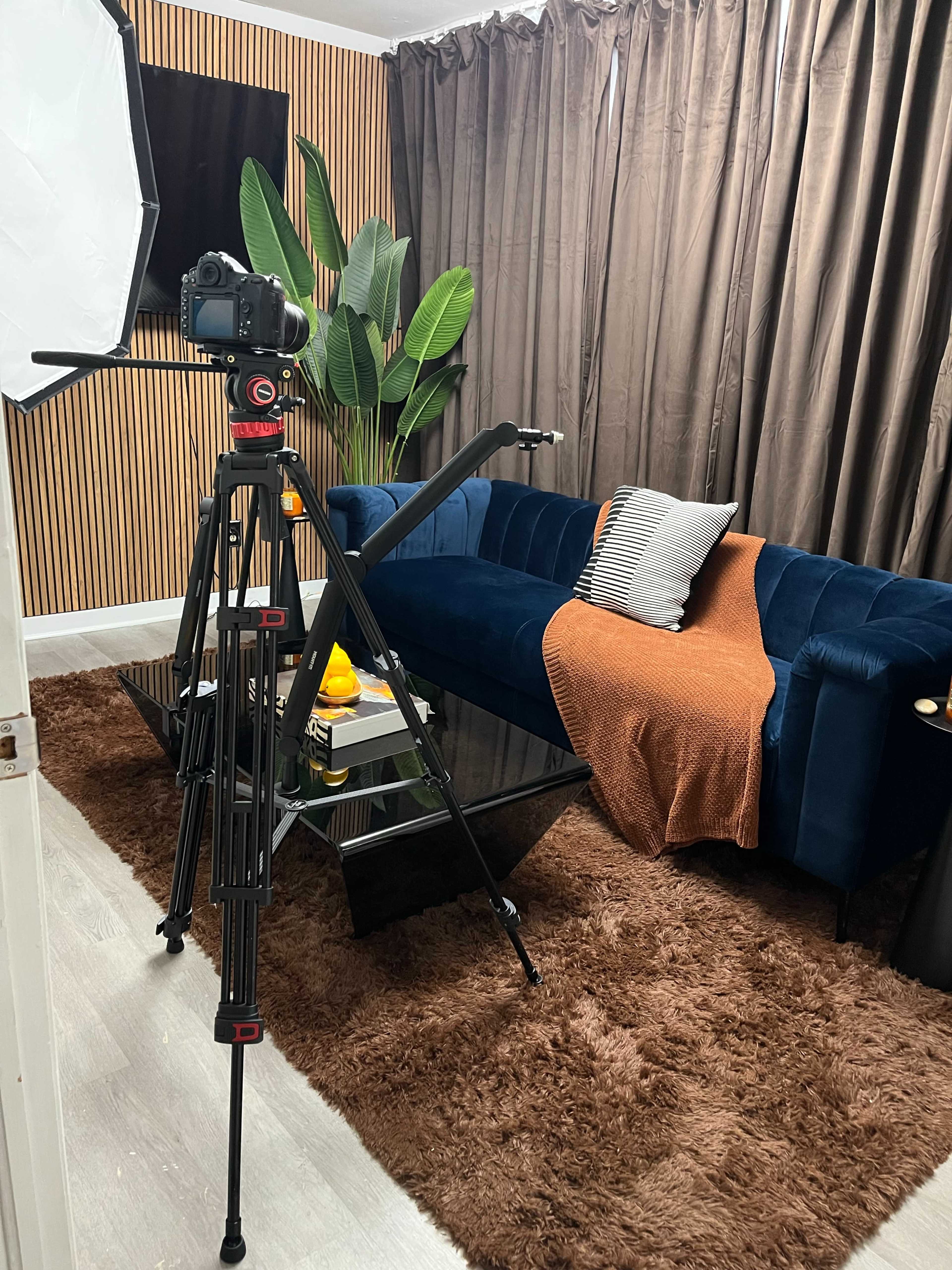 A camera on a tripod is positioned in front of a blue couch, with a blanket draped over it, in a room decorated with textured walls and curtains.