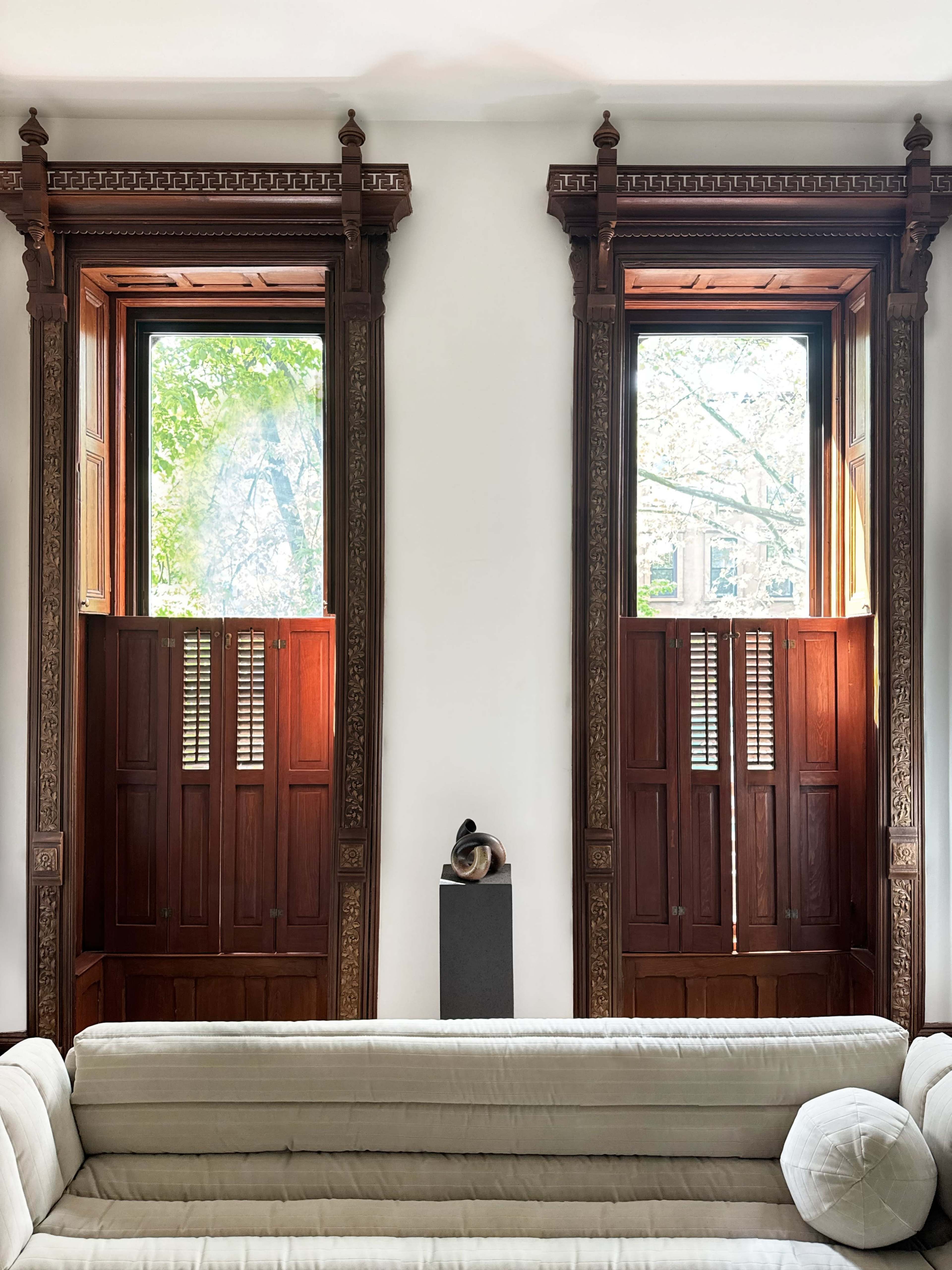 The image shows two ornate wooden windows on either side of a beige couch, with a sculpture on a pedestal in between them.