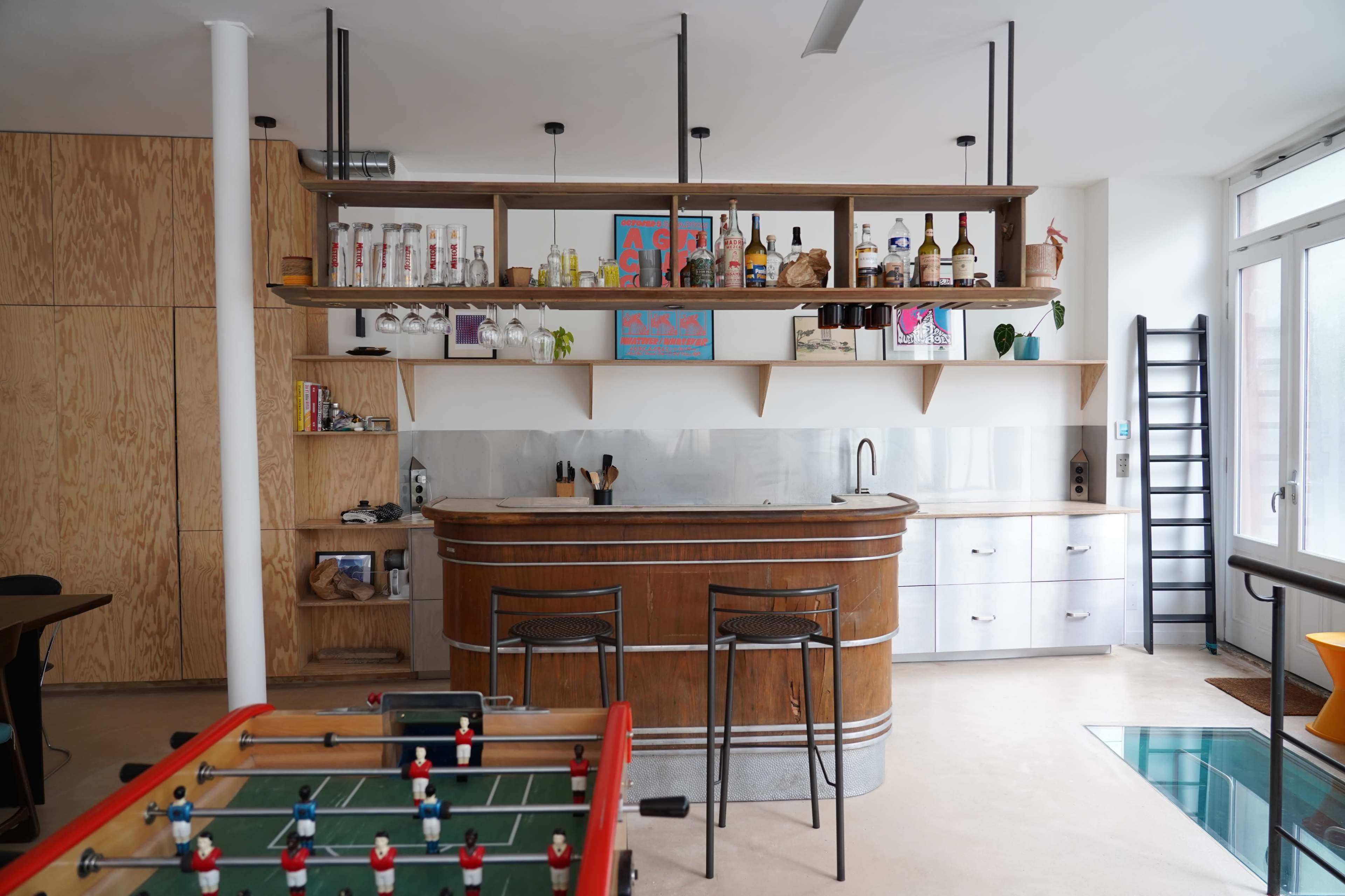 The scene features a modern kitchen and bar area with a wooden countertop, shelves displaying various bottles, and a foosball table in the foreground.