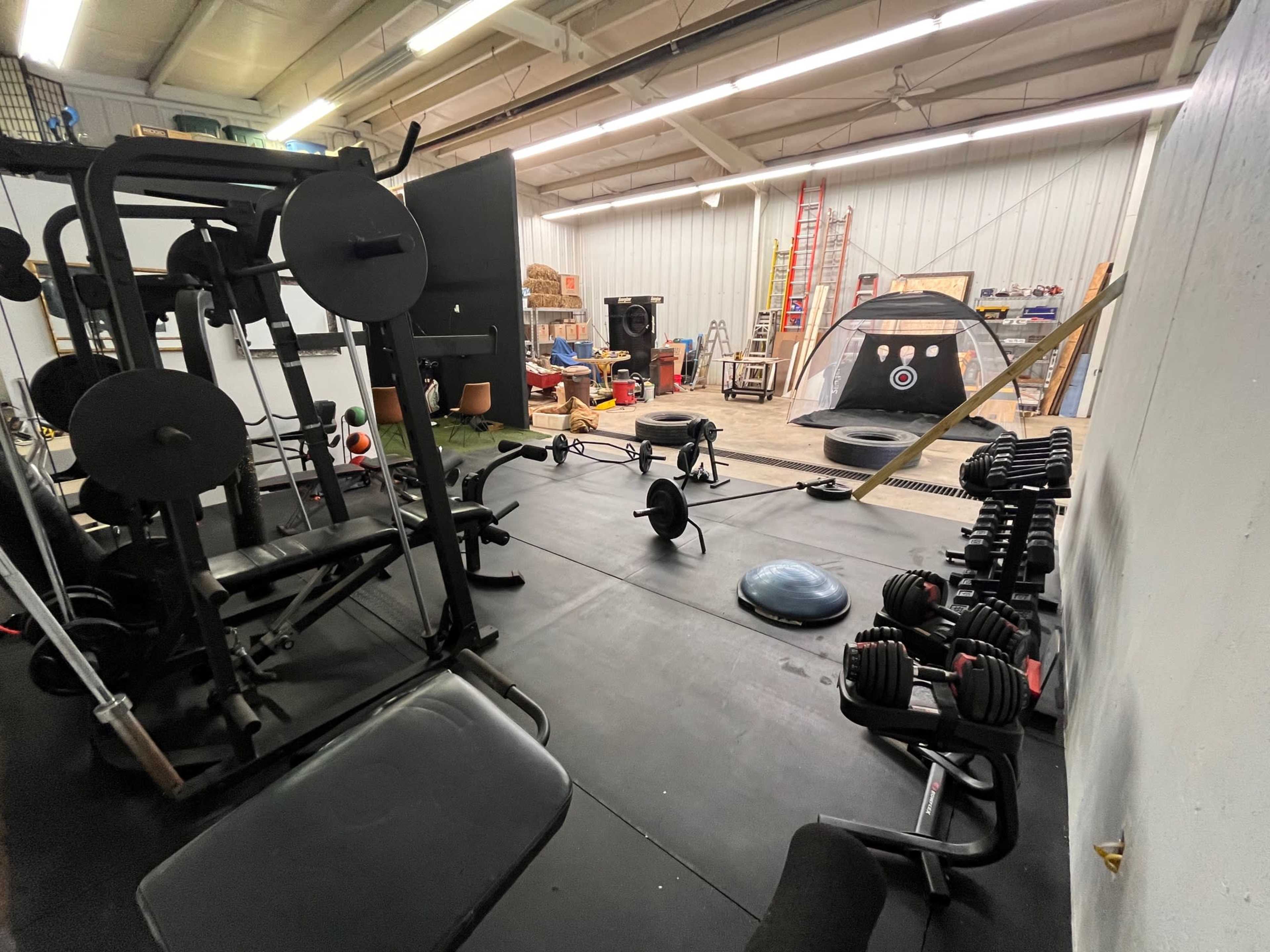 The image shows a gym space equipped with various weightlifting machines, free weights, and exercise equipment, alongside a ladder and a tent in the background.