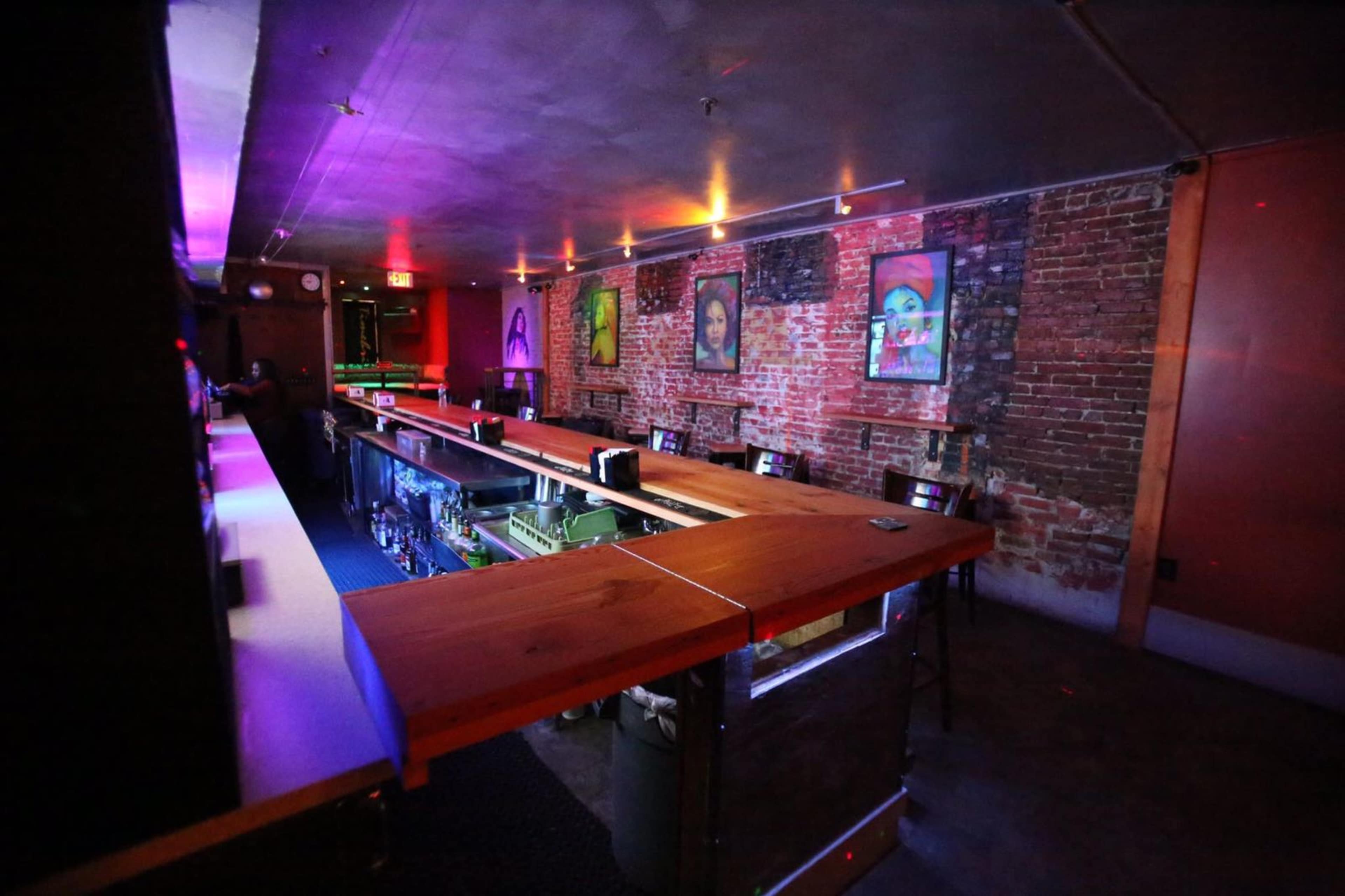A dimly lit bar with a wooden counter, exposed brick walls, and colorful artwork hanging on the walls.