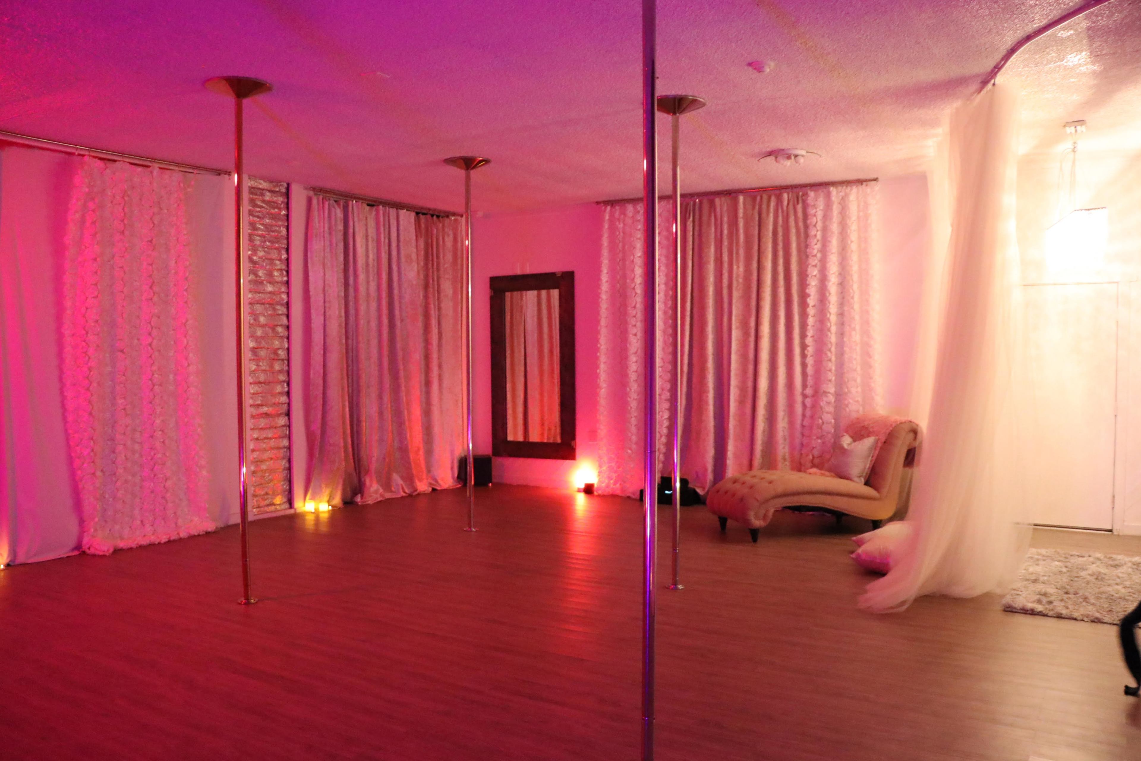A dance studio featuring polished wood floors, several pole dancing poles, and soft lighting against draped fabric along the walls.