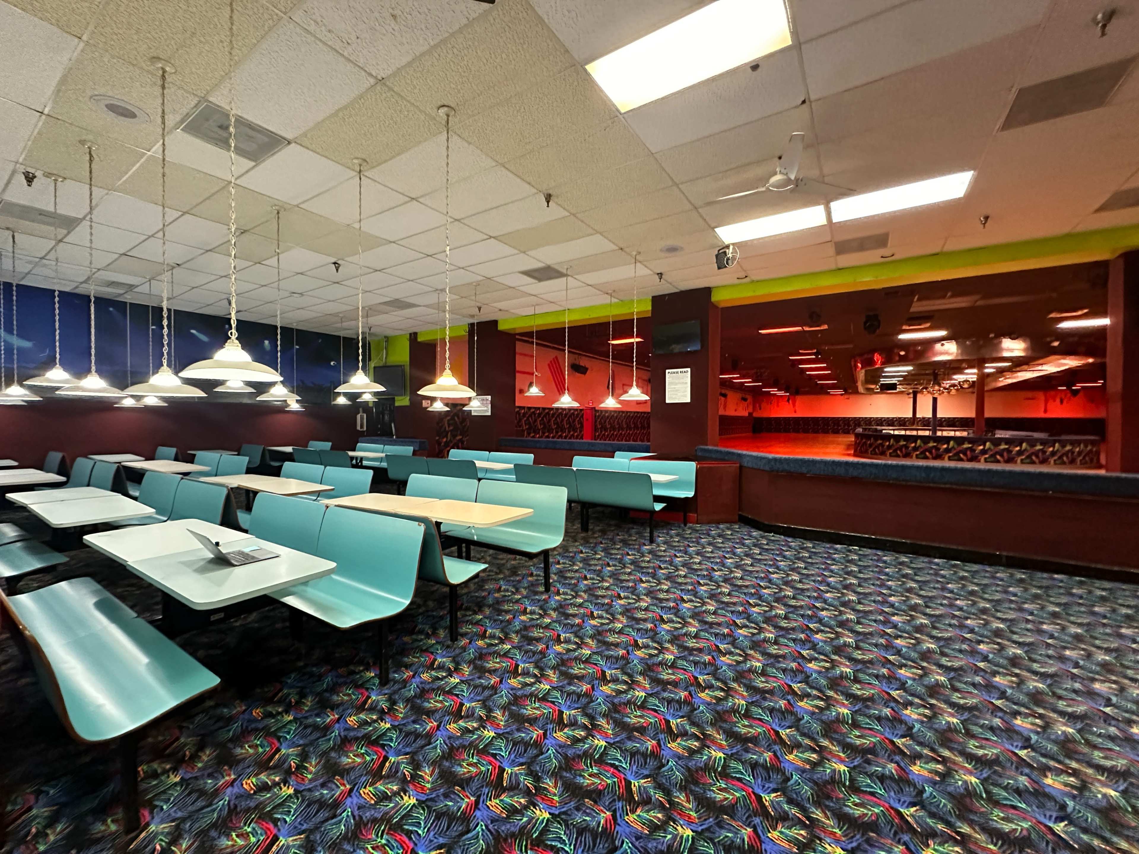 The image shows a large, retro-styled diner with teal booths and tables, featuring a colorful, patterned carpet and pendant lights hanging from the ceiling.